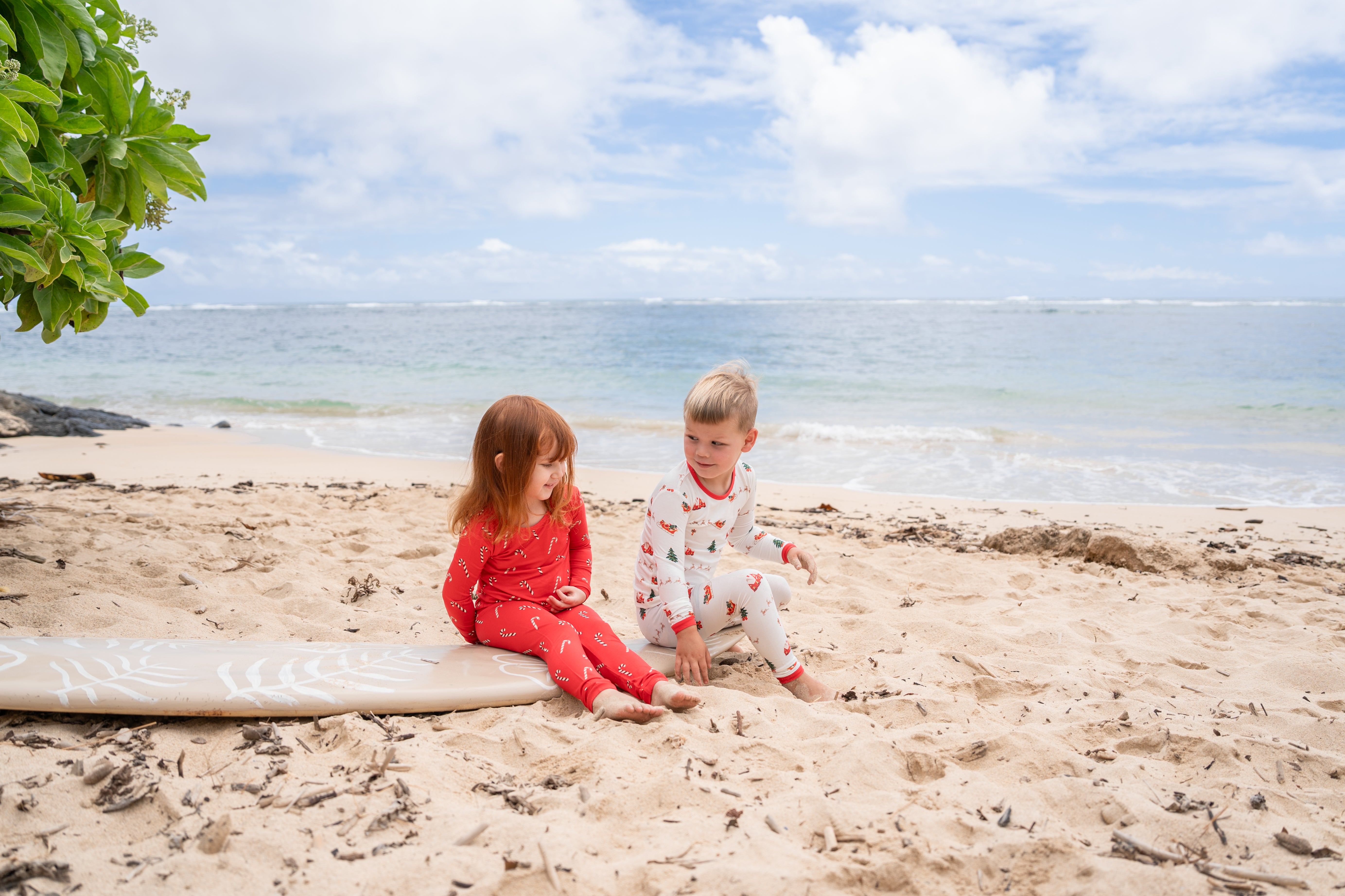 2 kids wearing christmas kyte baby pajamas sitting on a surf board on the beach