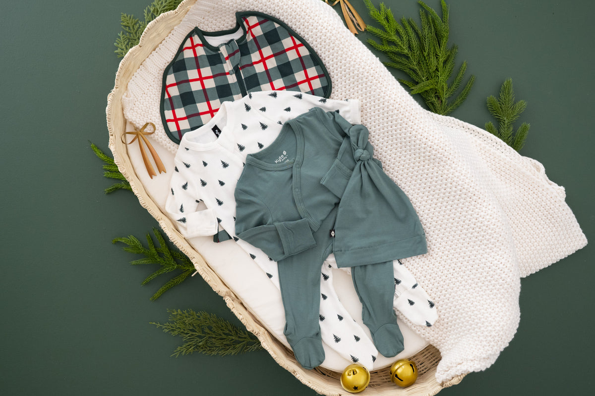 A styled flat-lay of baby clothing arranged in a woven basket with a white knit blanket, green pine branches, and small gold bells on a dark green background. The garments include a plaid bib, a white long-sleeve top with small christmas tree motifs, a muted green romper
