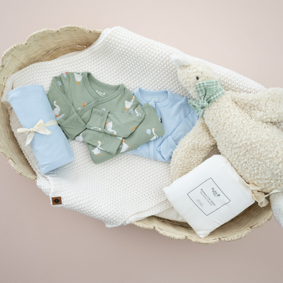 Flat lay image of newborn essentials including a crib sheet, silly goose outfit, light blue swaddle and footie and stuffed goose, in a wicker bassinet