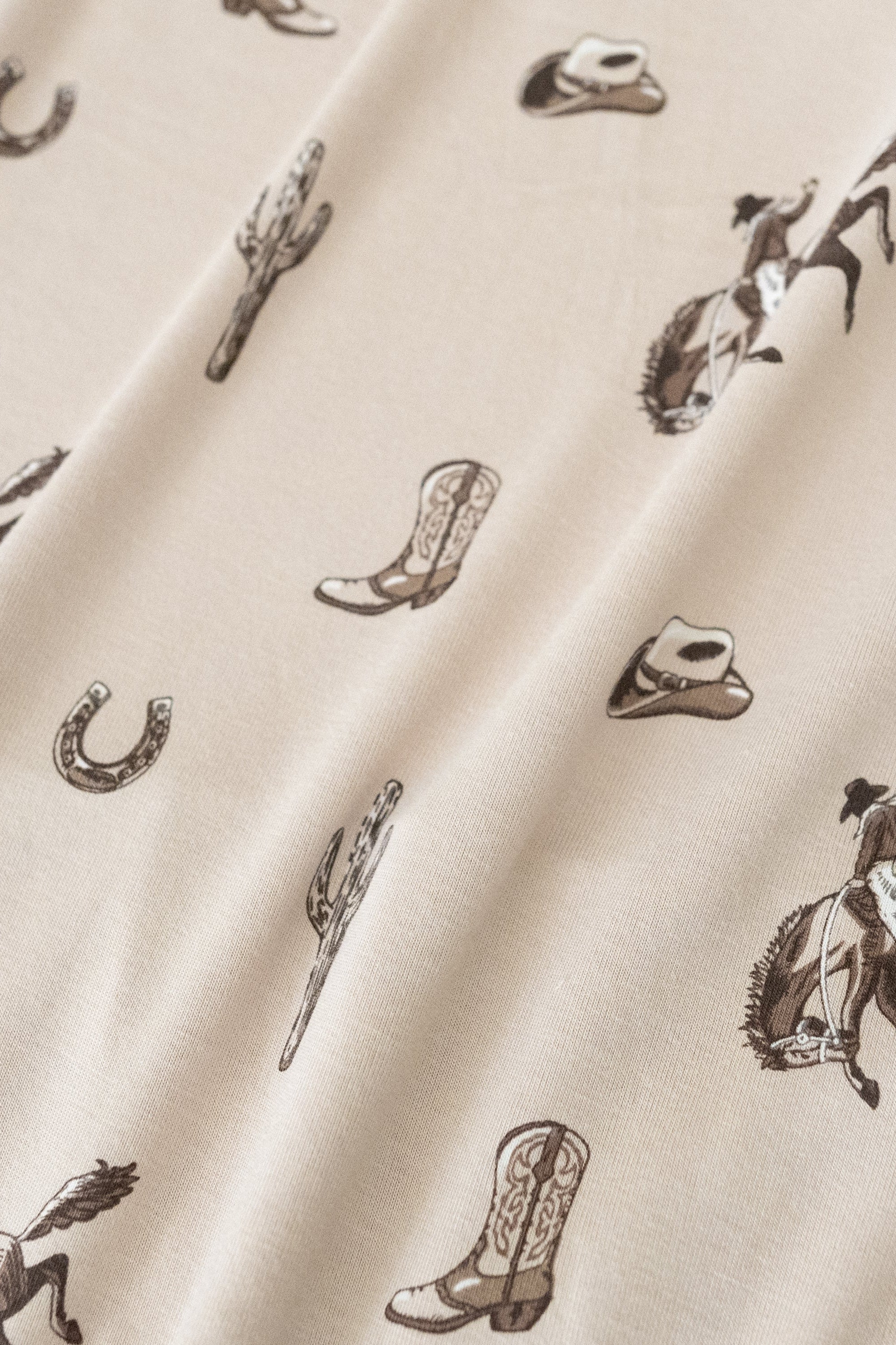Close up of bamboo fabric: A repeating, all-over print in monochromatic browns, featuring: a cowboy riding a bucking horse; a western boot: a three-arm cactus; a horseshoe; and a cowboy hat, over a dusty beige color named Bisque.