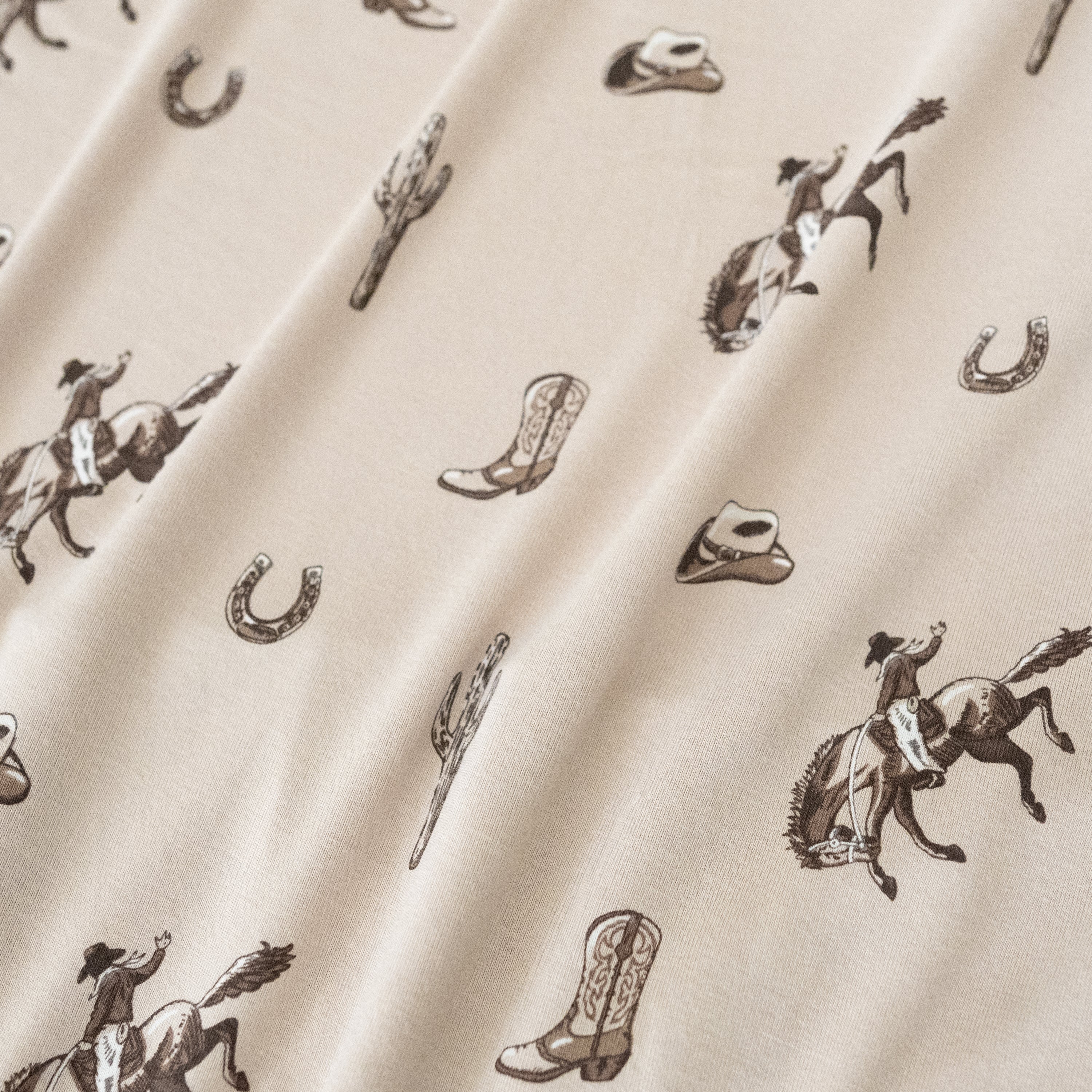 Close up of bamboo fabric: A repeating, all-over print in monochromatic browns, featuring: a cowboy riding a bucking horse; a western boot: a three-arm cactus; a horseshoe; and a cowboy hat, over a dusty beige color named Bisque.