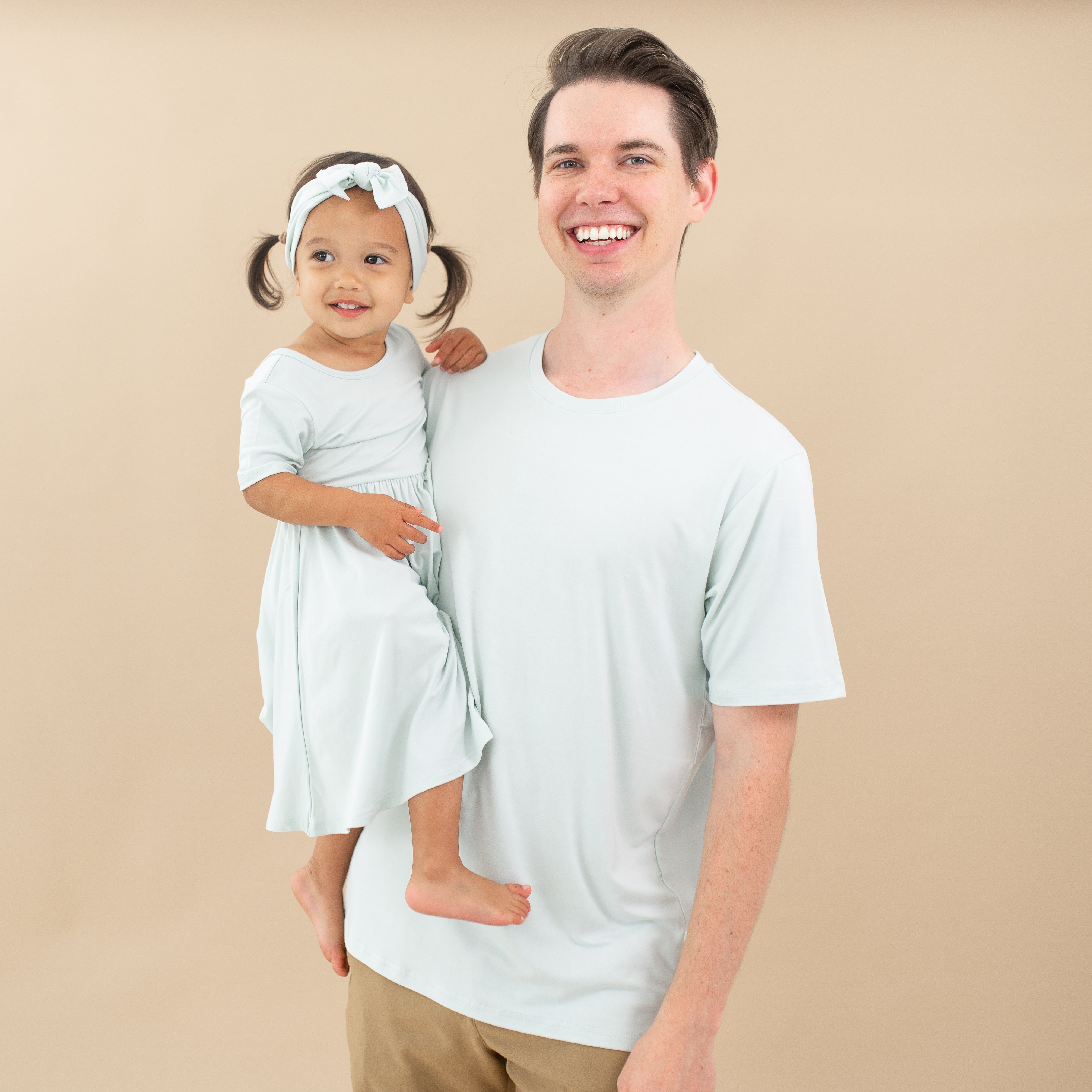 Male model wearing the Men's Crew Neck Tee in Dew holding his daughter who is in a Twirl Dress in Dew
