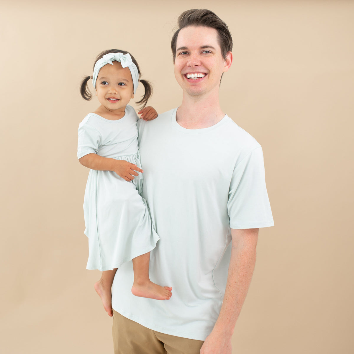 Male model wearing the Men's Crew Neck Tee in Dew holding his daughter who is in a Twirl Dress in Dew