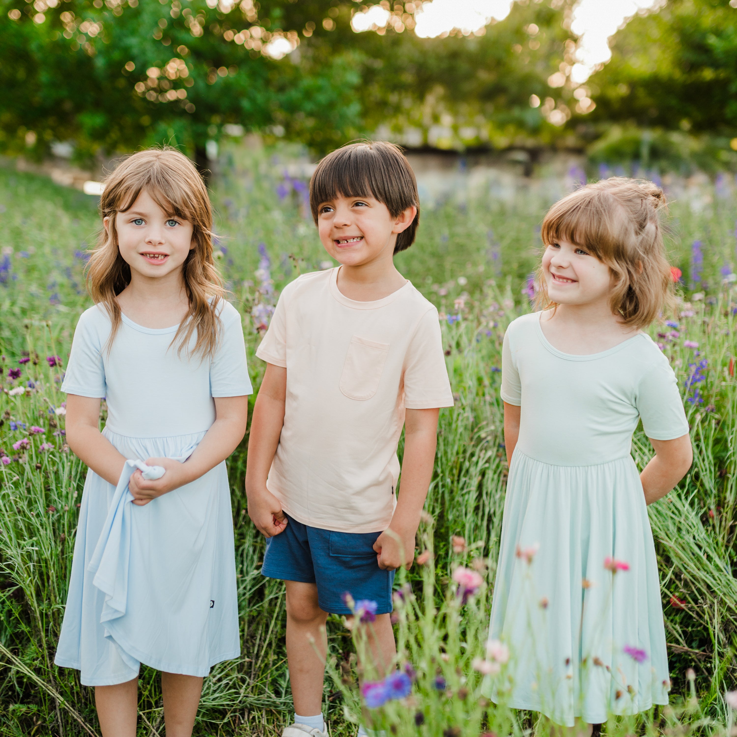 Three children models standing in wild flowers outdoors. Models are wearing a Twirl Dress in Mist, Canyon Crew Neck tee with Blue shorts and a Twirl Dress in Dew