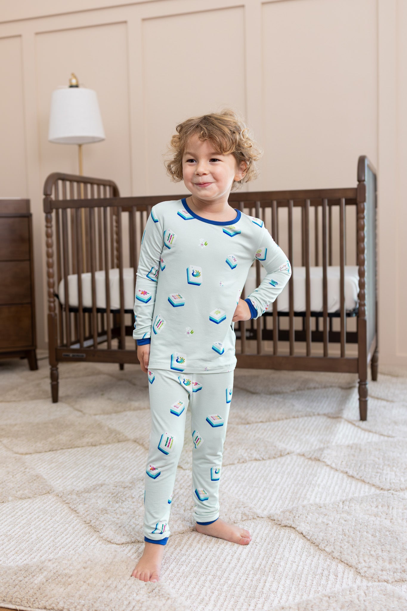 Boy wearing Long Sleeve Pajamas in Dew Mahjong print posing in front of a crib