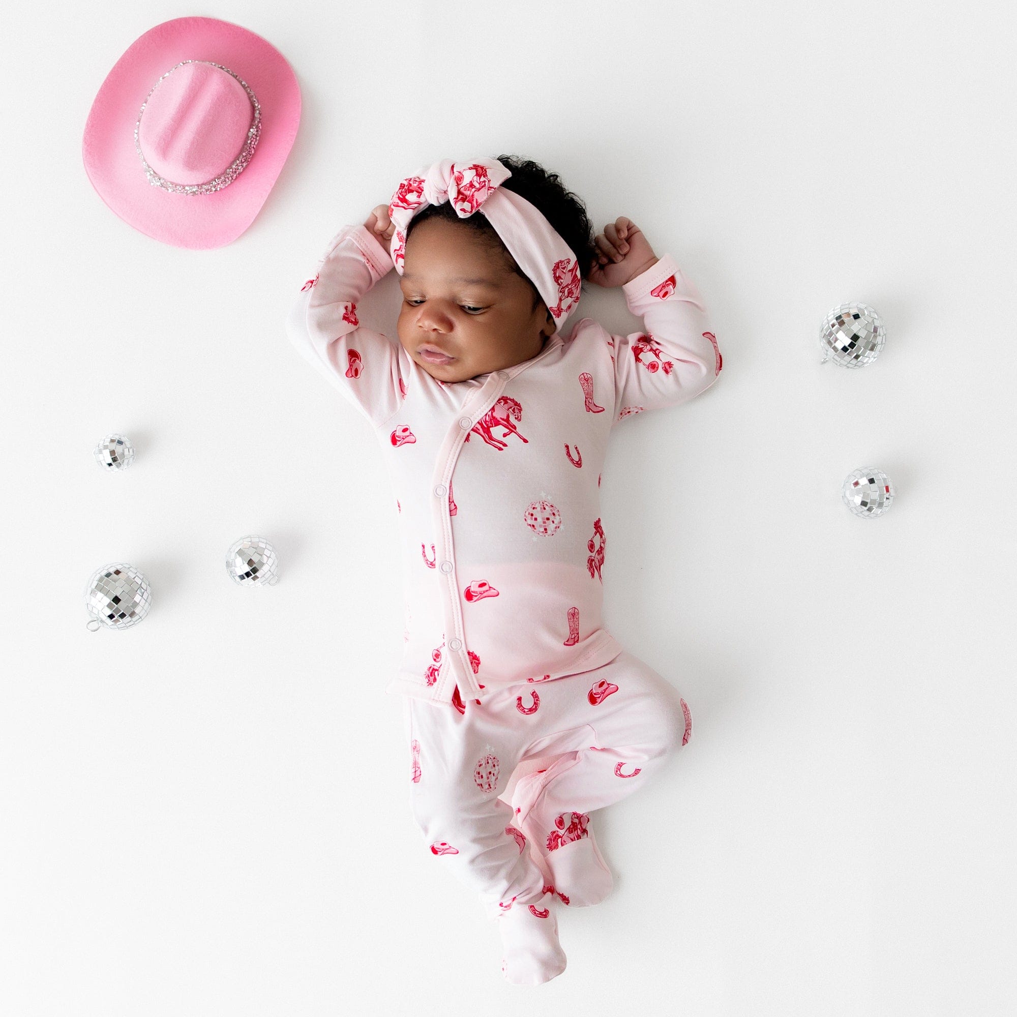 Infant girl laying on a light cream surface wearing the breathable Take Me Home Set with Bow in Disco Cowgirl from Kyte Baby with toy disco balls and a pink cowboy hat around her