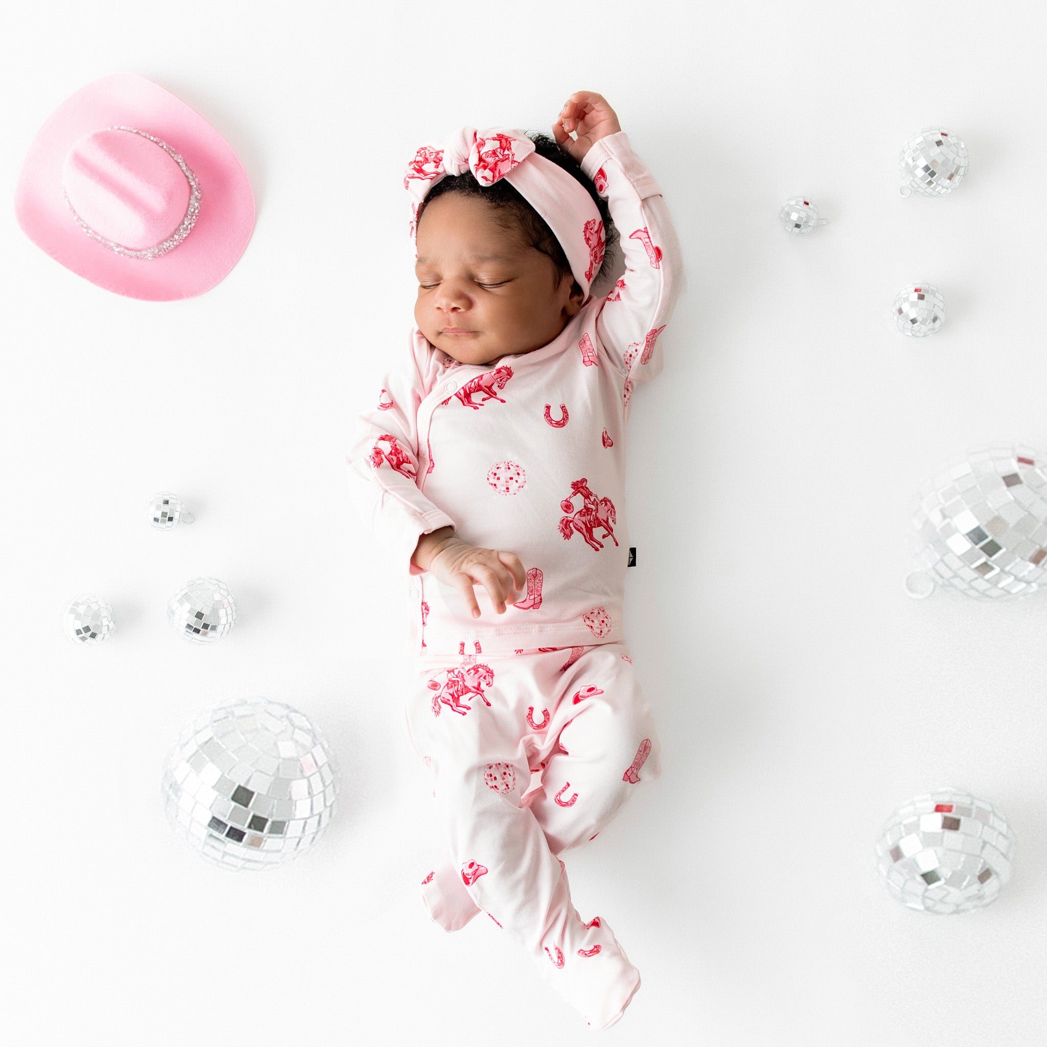 Newborn laying on a cream surface stretching wearing the breathable Take Me Home Set with Bow in Disco Cowgirl from Kyte Baby surrounded by toy disco balls and pink cowboy hat