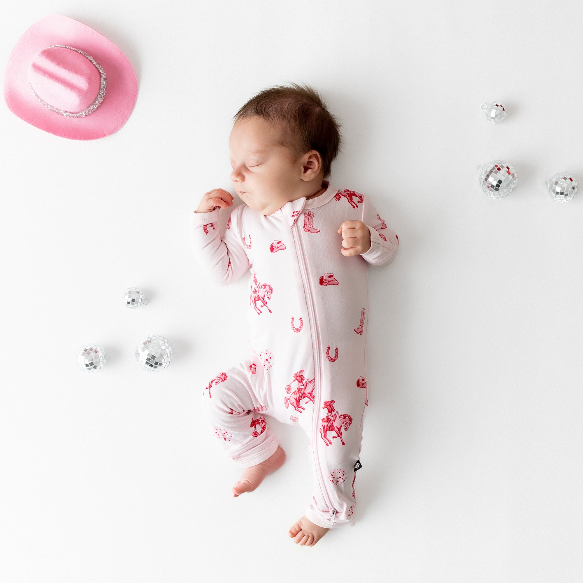 Newborn laying on a light cream neutral surface wearing the breathable Zippered Romper in Disco Cowgirl from Kyte Baby with toy disco ball props and pink cowboy hat around her
