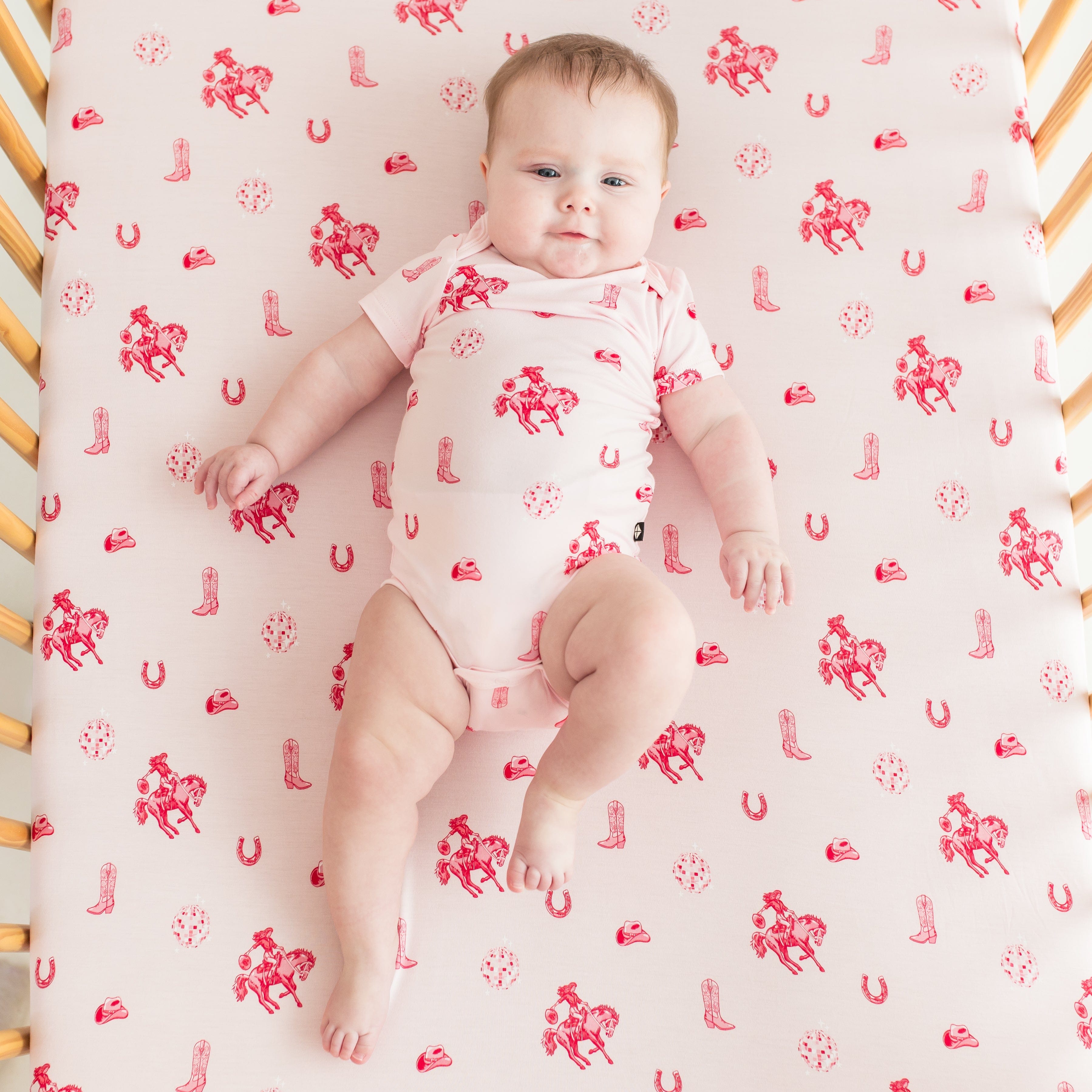 Infant girl wearing the Bodysuit in Disco Cowgirl laying in a crib on a matching crib sheet