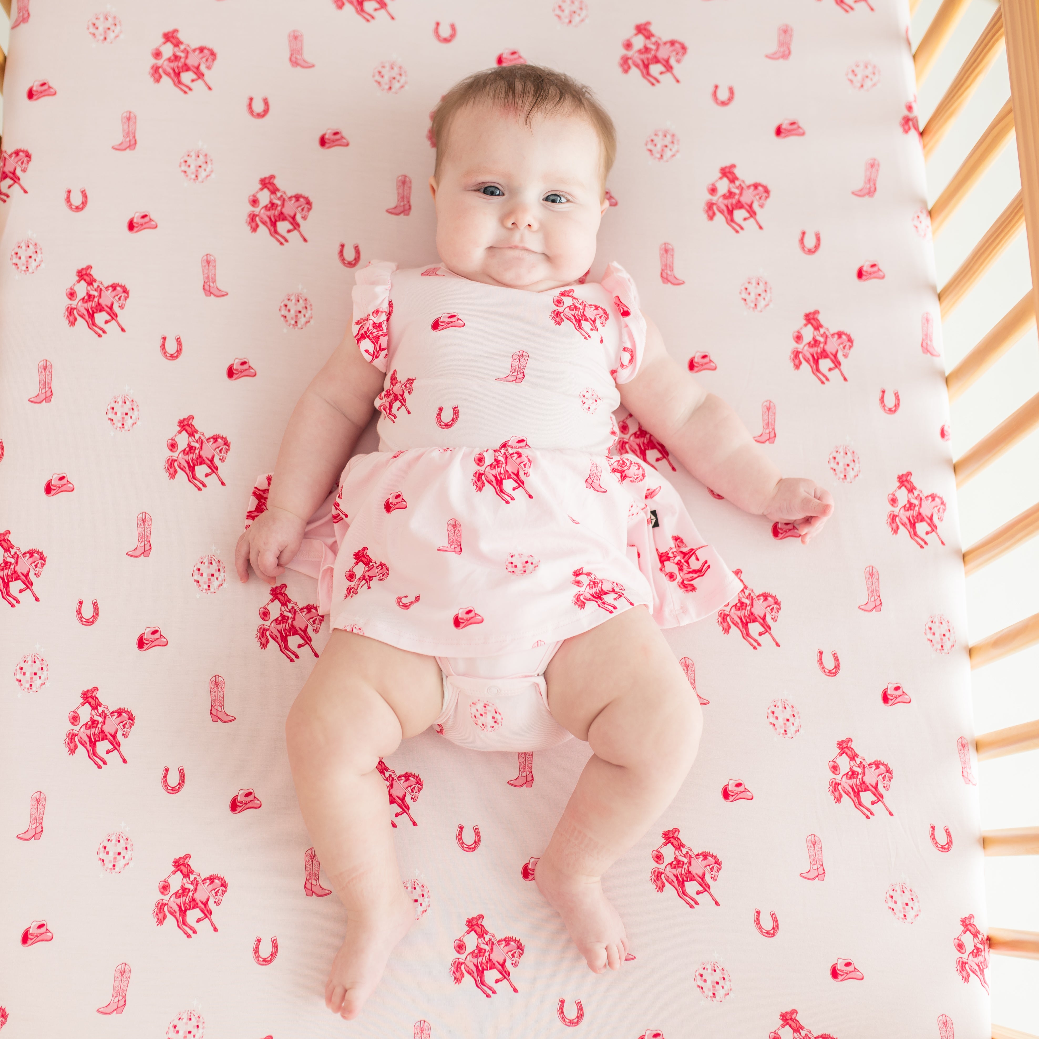 Young infant girl wearing the Twirl Bodysuit Dress in Disco Cowgirl laying in a crib on a matching crib sheet