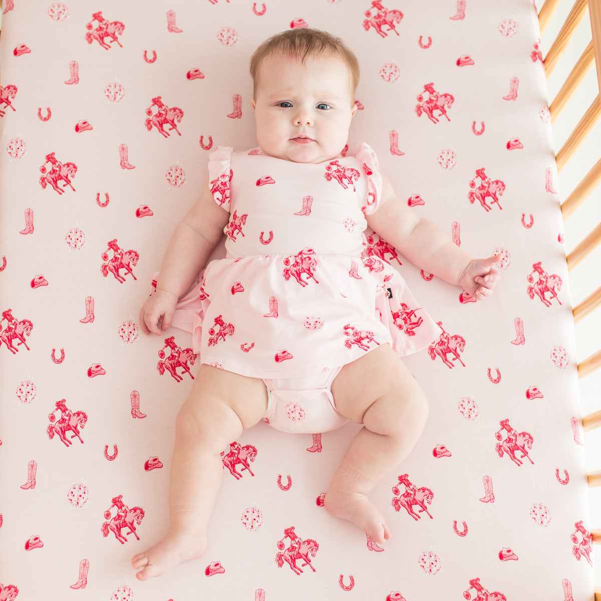 Infant girl laying in a crib on the Disco Cowgirl crib sheet modeling the matching short sleeve bodysuit twirl dress
