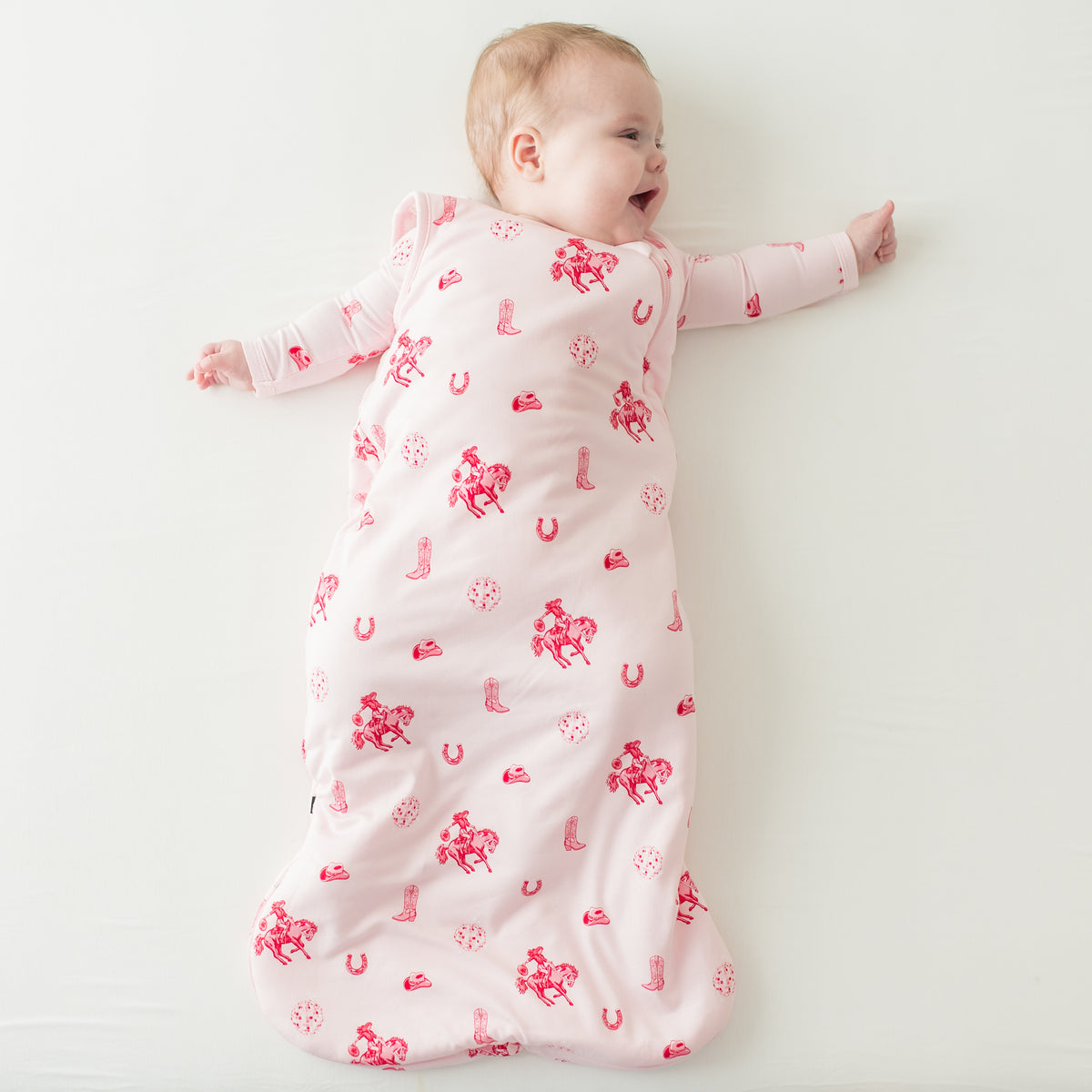 Young infant girl laying on a cream surface wearing the Sleep Bag in Disco Cowgirl 1.0 and matching long sleeve romper
