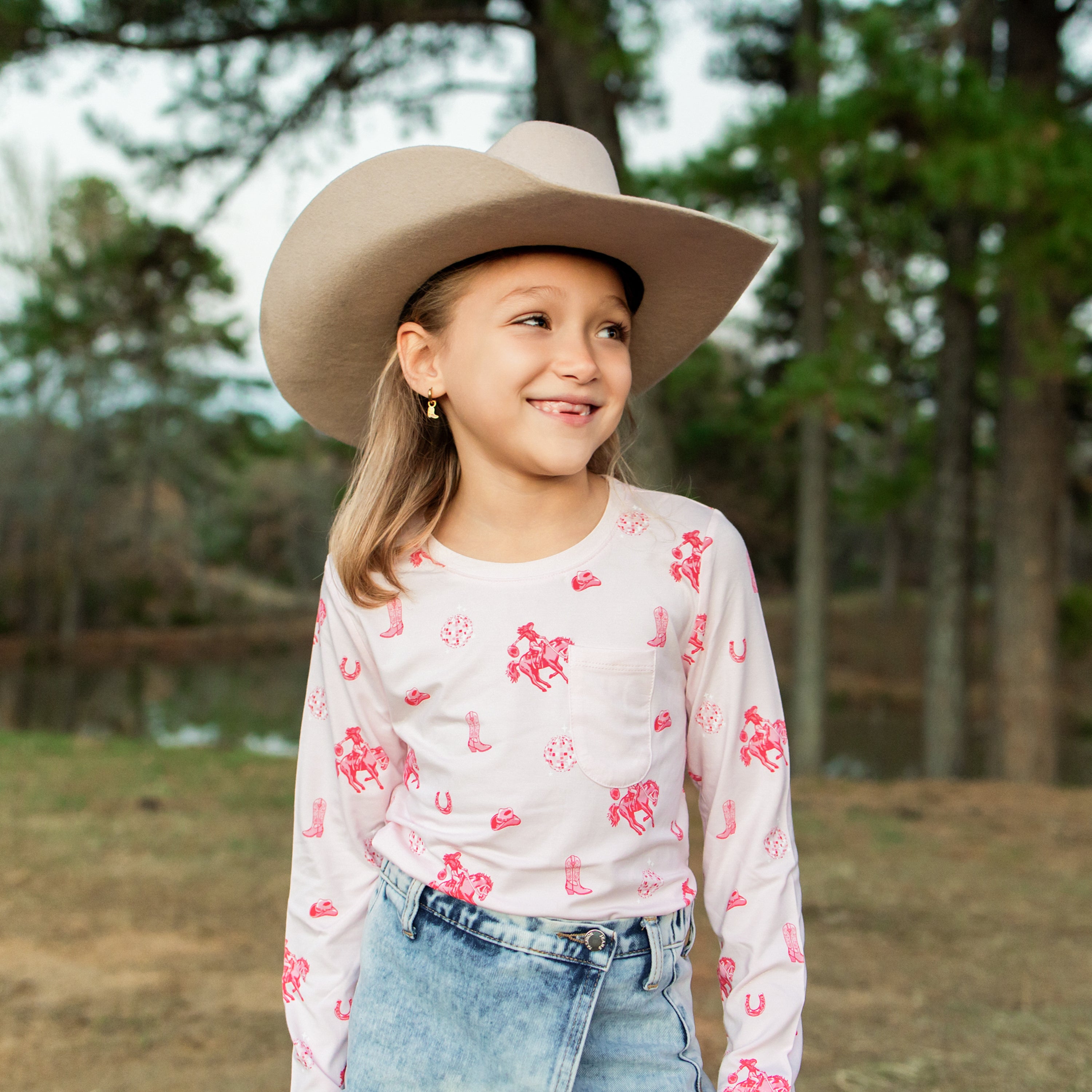Young girl outside in front of trees wearing the Long Sleeve Toddler Crew Neck Tee in Disco Cowgirl with a denim skort and western hat