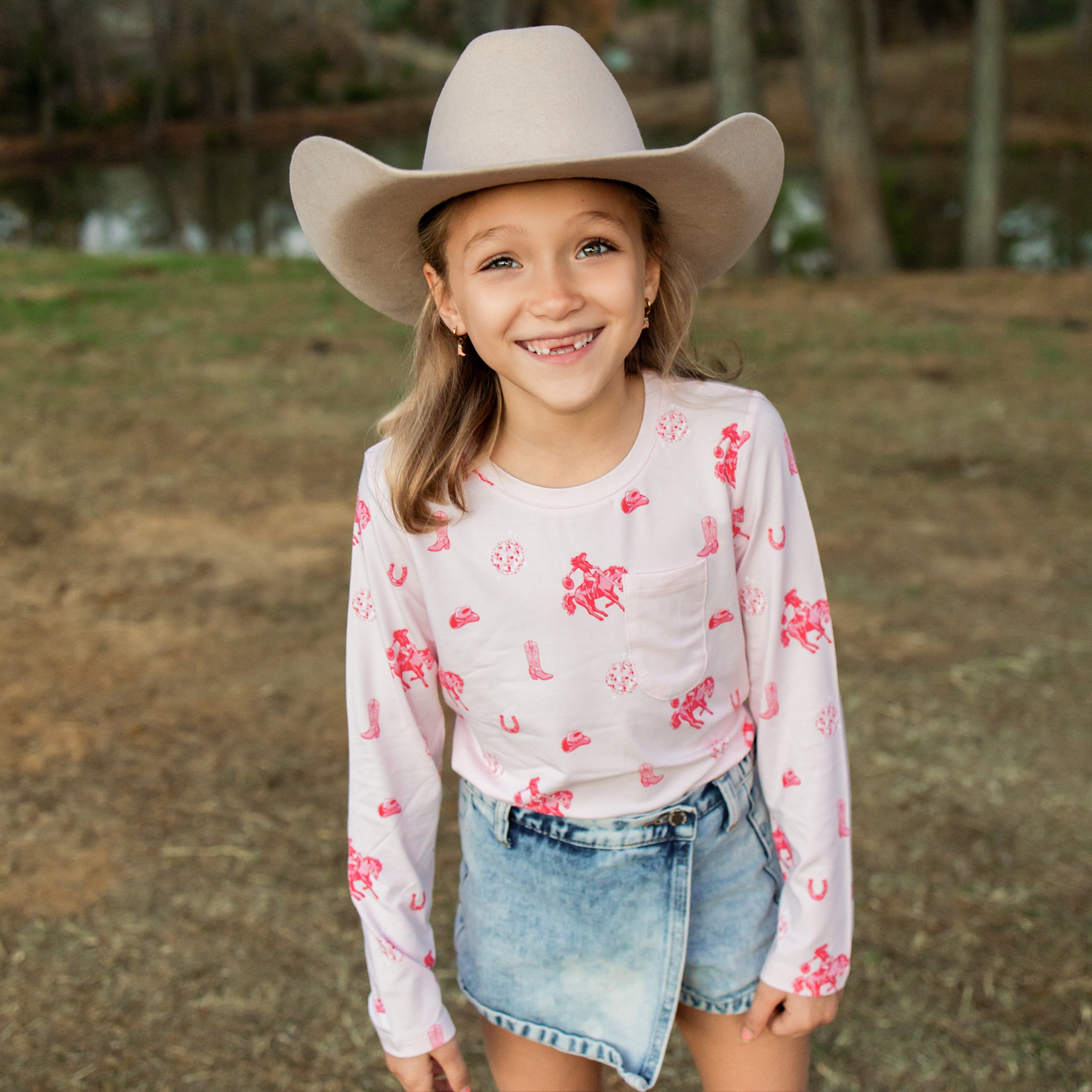 Smiling girl standing in a field in front of trees wearing the Long Sleeve Toddler Crew Neck Tee in Disco Cowgirl with a denim skort and light neutral western hat