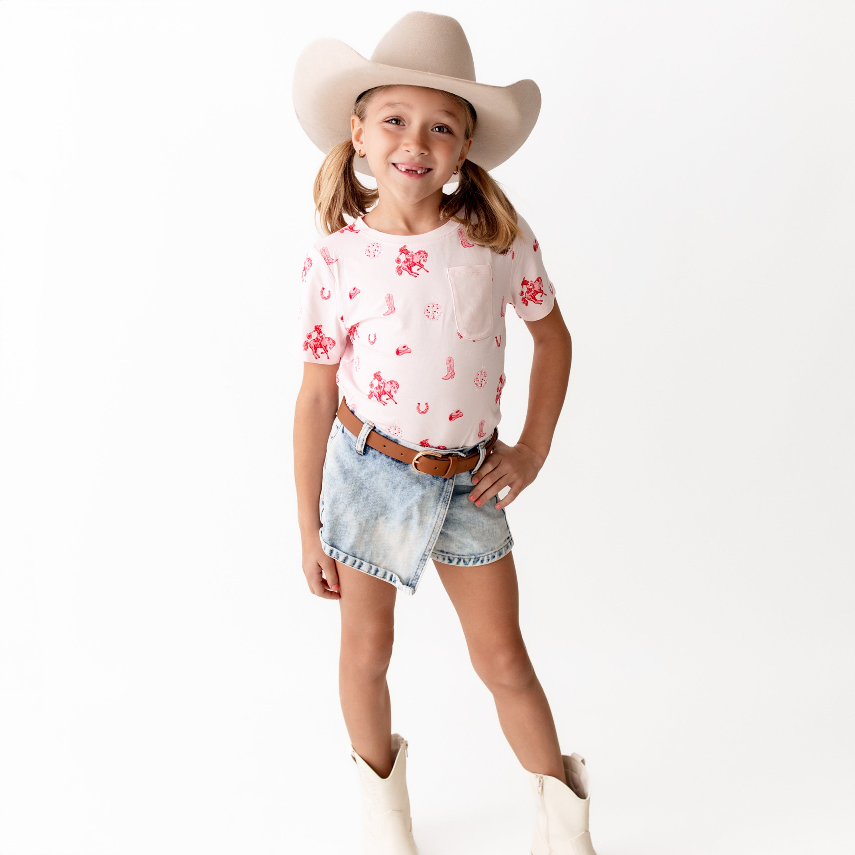 Young girl standing in front of a white colored background wearing the Toddler Crew Neck Tee in Disco Cowgirl paired with a jean skort, white cowboy boots and a light colored cowboy hat