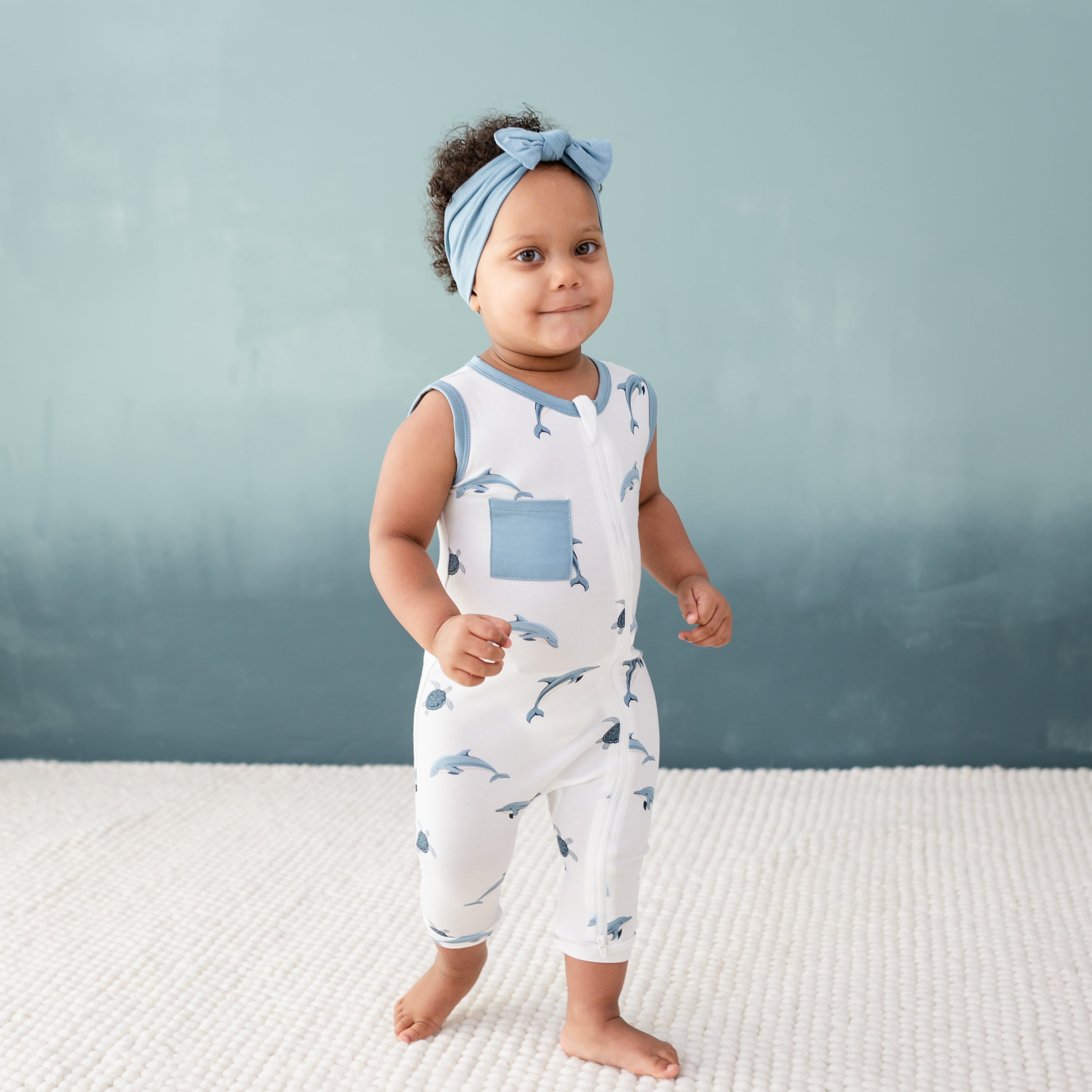 Toddler wearing Kyte Baby Zippered Sleeveless Romper in Dolphin with Dusty Blue Bow