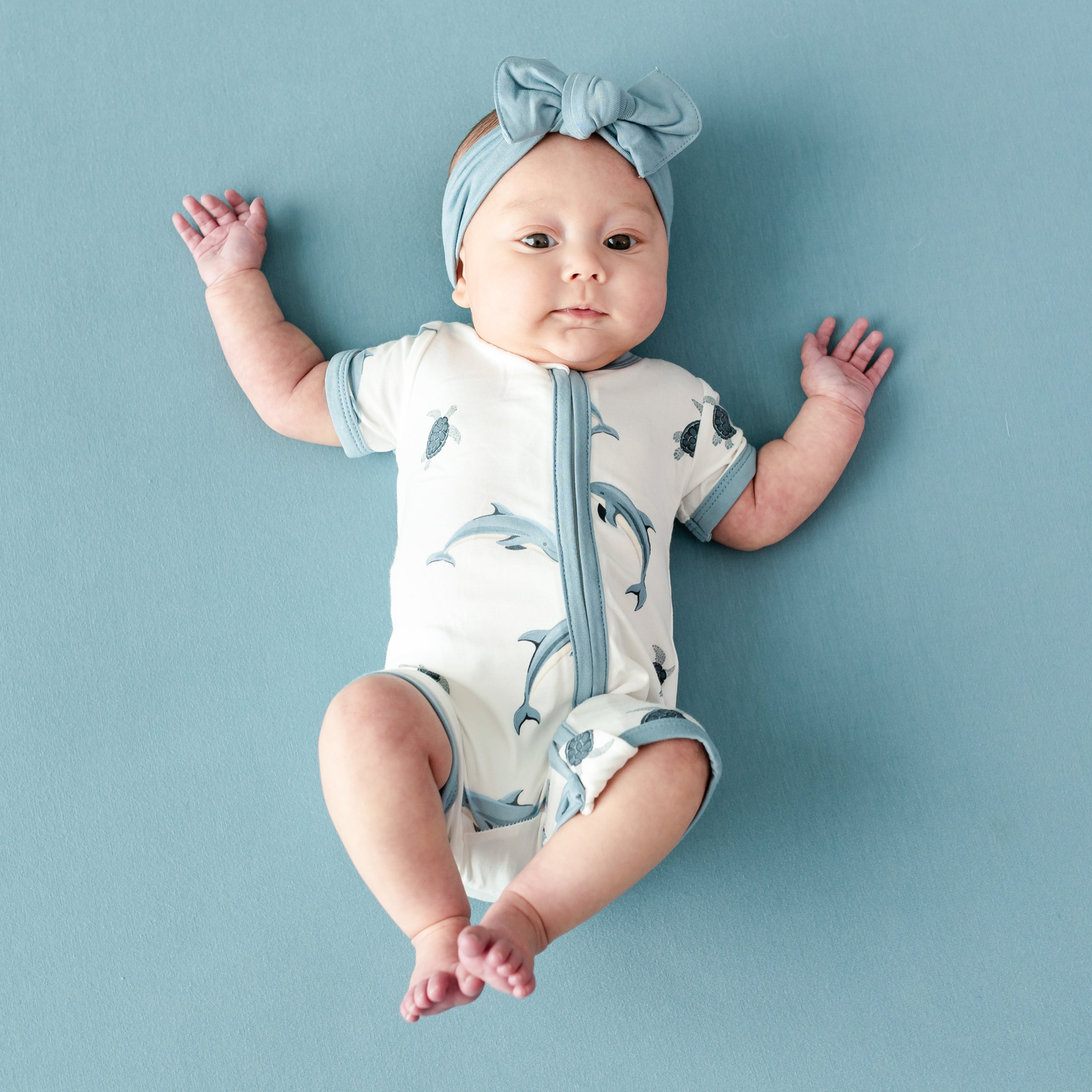 Infant wearing Kyte Baby Zippered Shortall Romper in Dolphin with Dusty Blue Bow