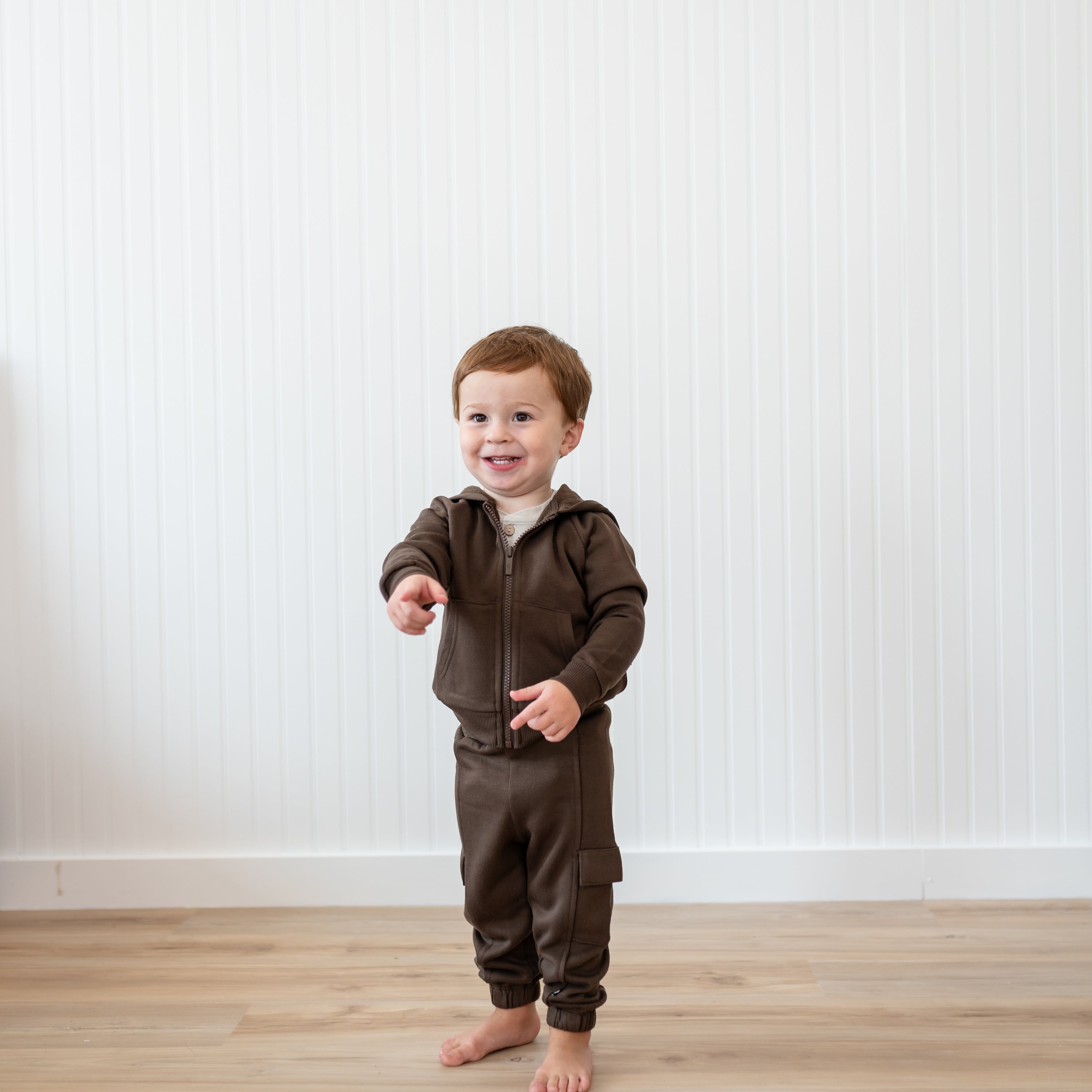 Young boy standing in front of a white paneled wall wearing the Fleece Zip Up Hoodie in Espresso and matching fleece cargo pants pointing forwards