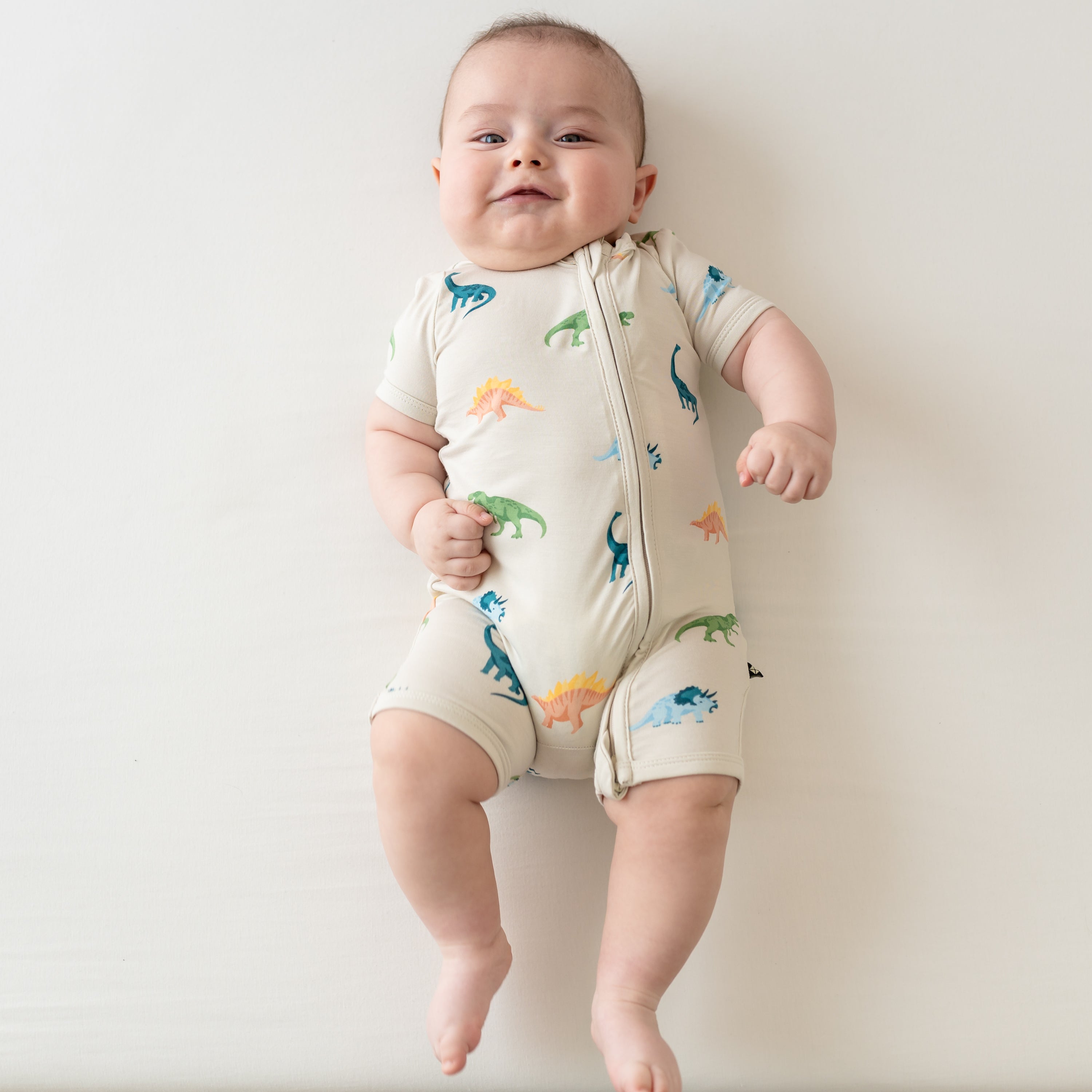 Infant wearing Kyte Baby Zippered Shortall Romper in Ecru Roar with dinosaurs