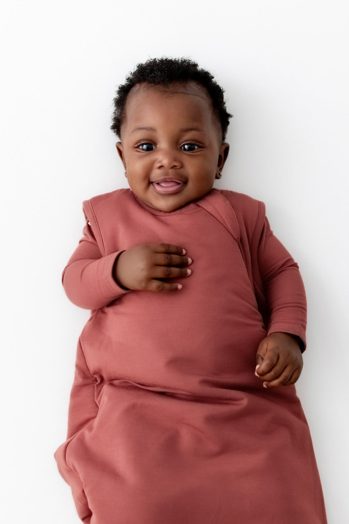 A smiling baby lying on their back, wearing a dusty terracotta 2.5 TOG sleep sack against a clean white background.