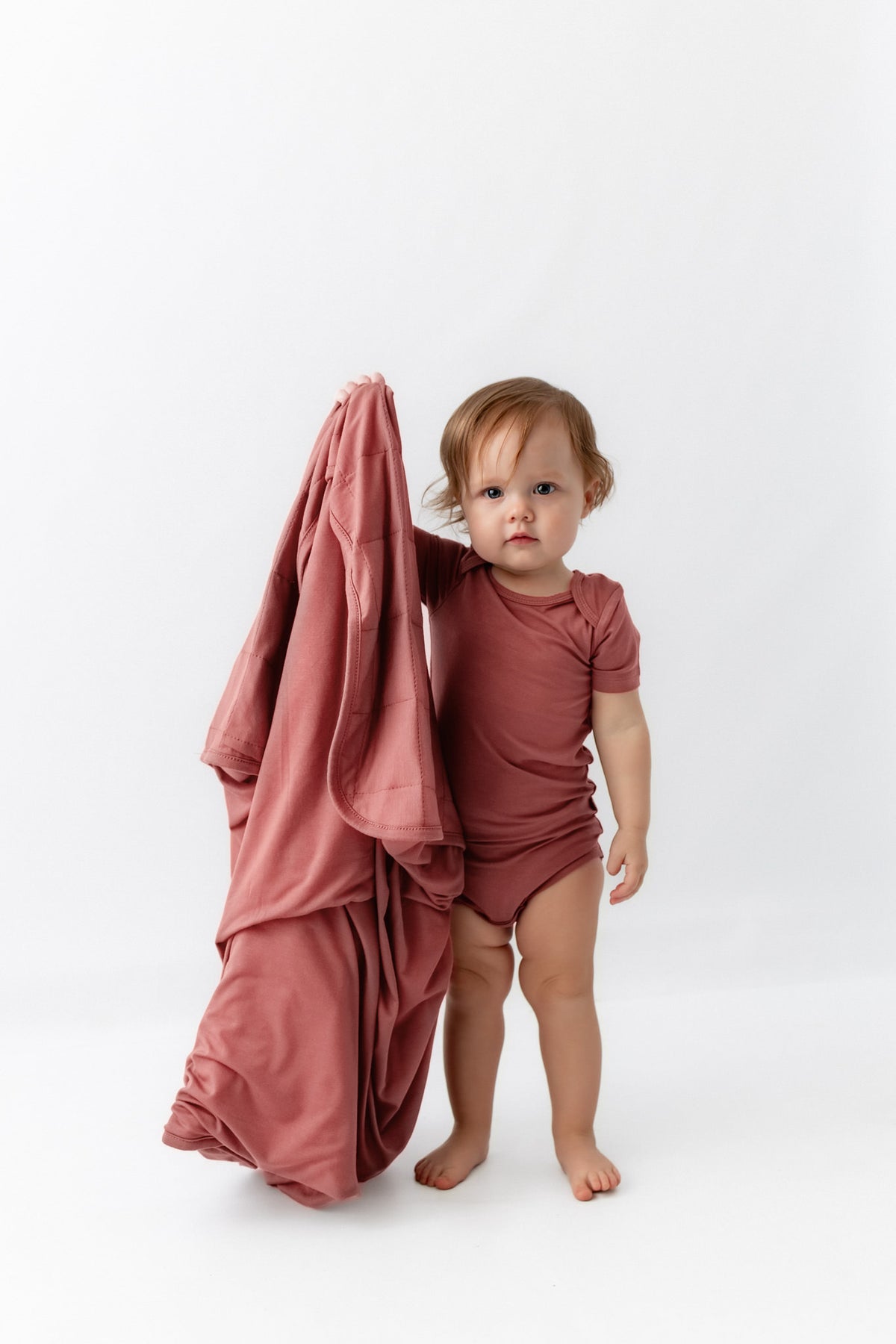 Baby holding Baby Blanket in Ember while wearing a similar color bodysuit dress on a white background