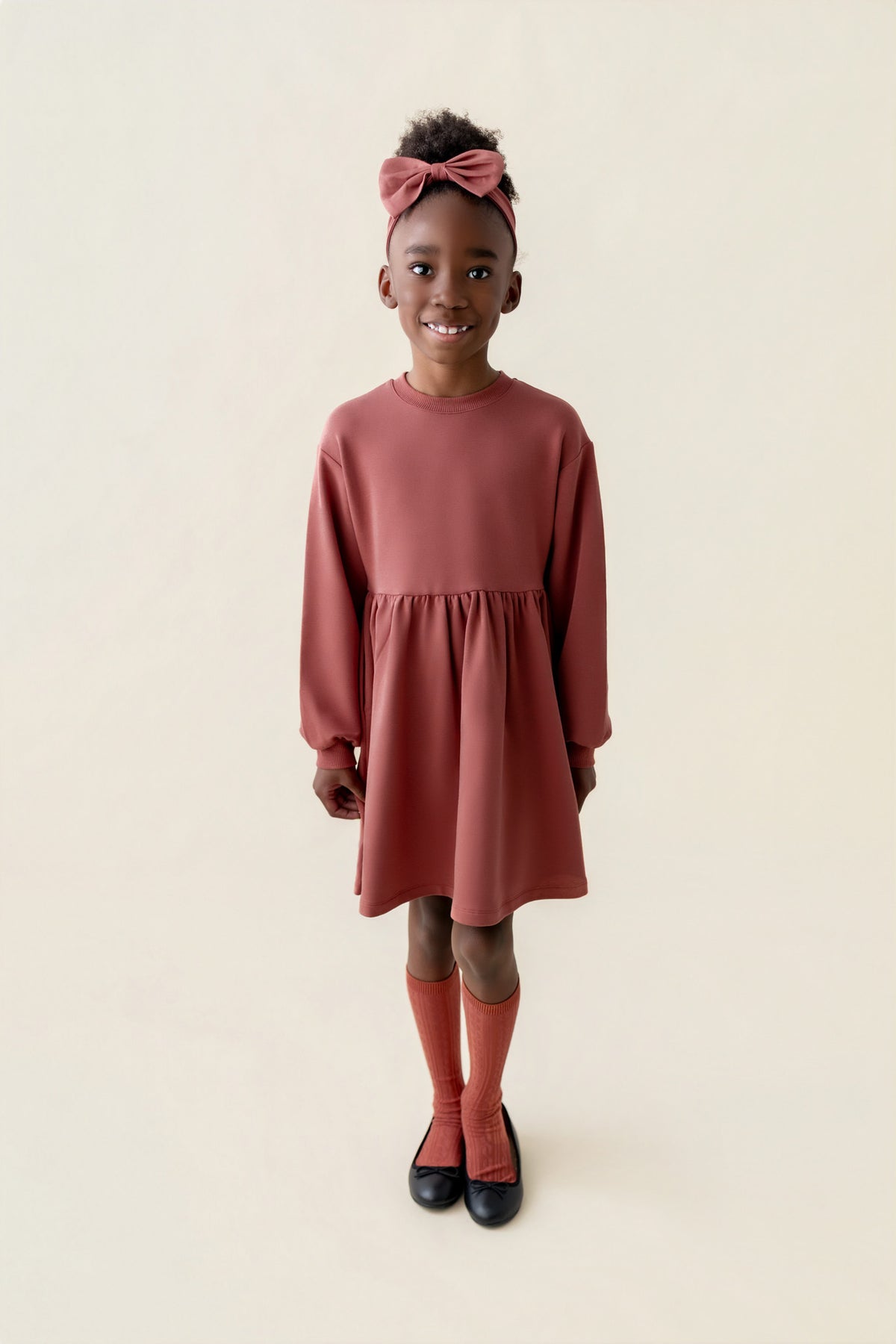Girl wearing Long Sleeve Skater Dress in Ember color with knotted bow headband on a beige background