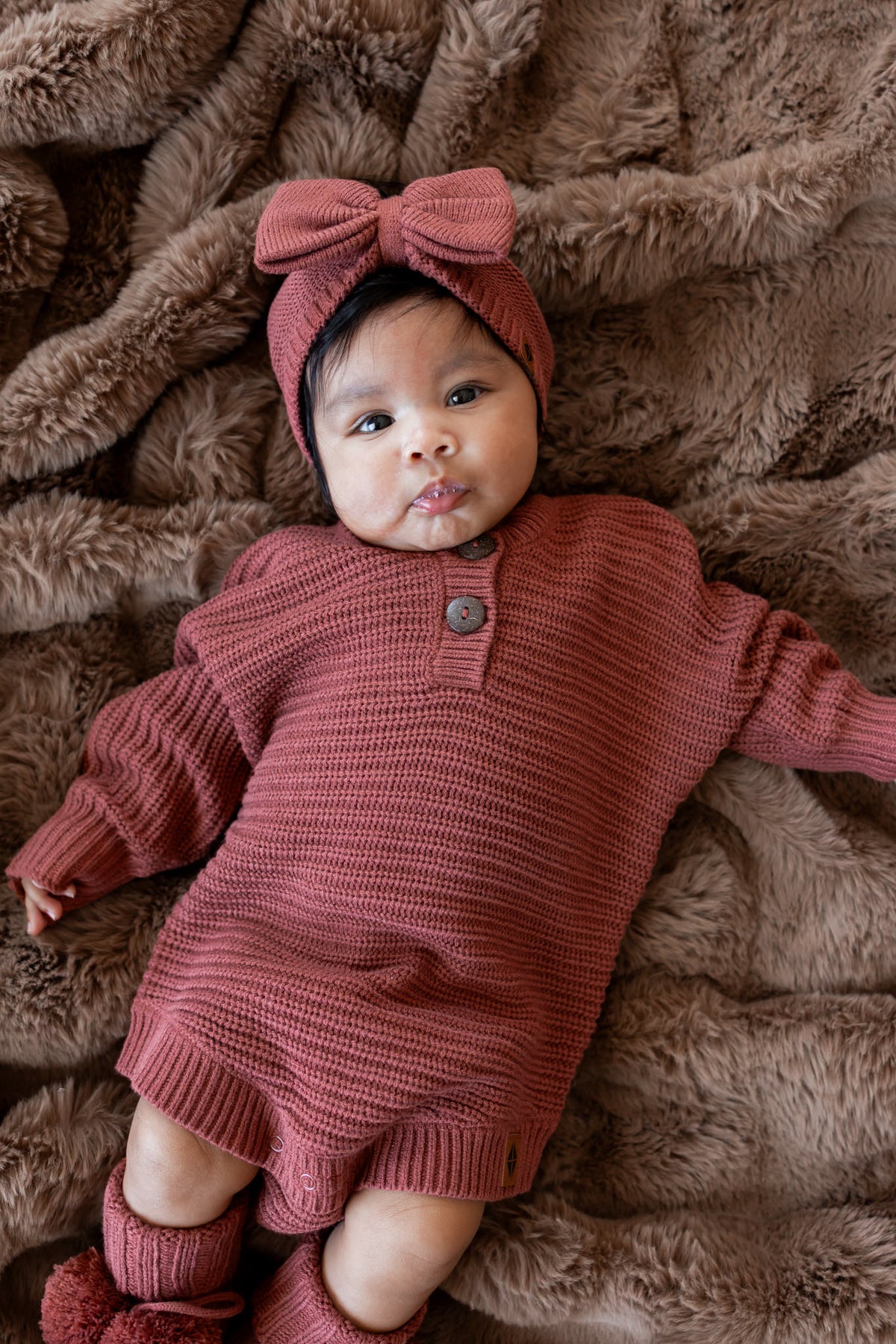 Baby lying on a brown blanket wearing Chunky Knit Romper, chunky knit bow headband and chunky knit booties in Ember