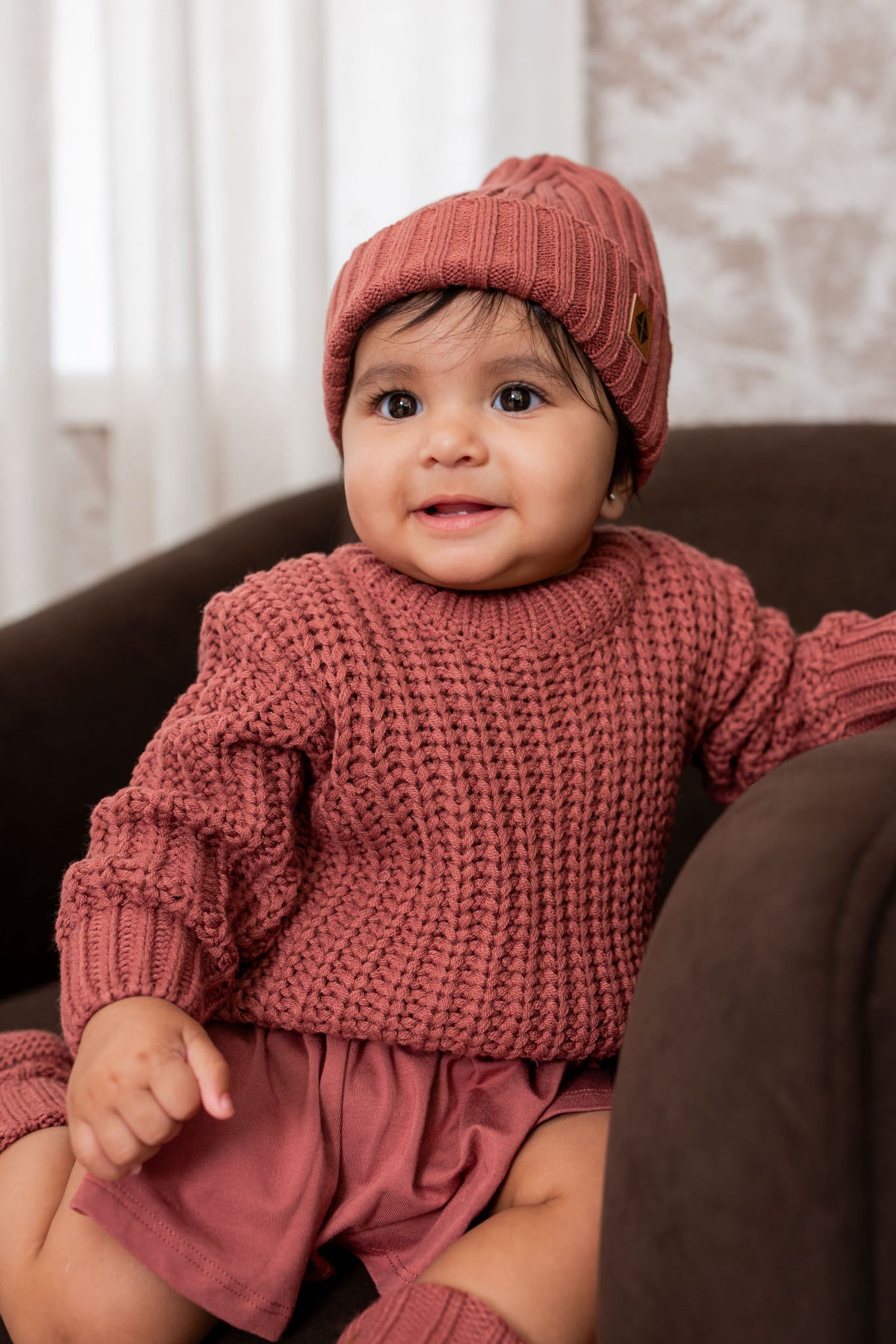 Baby wearing Chunky Knit Sweater and chunky knit ribbed beanie in Ember while sitting on a brown sofa