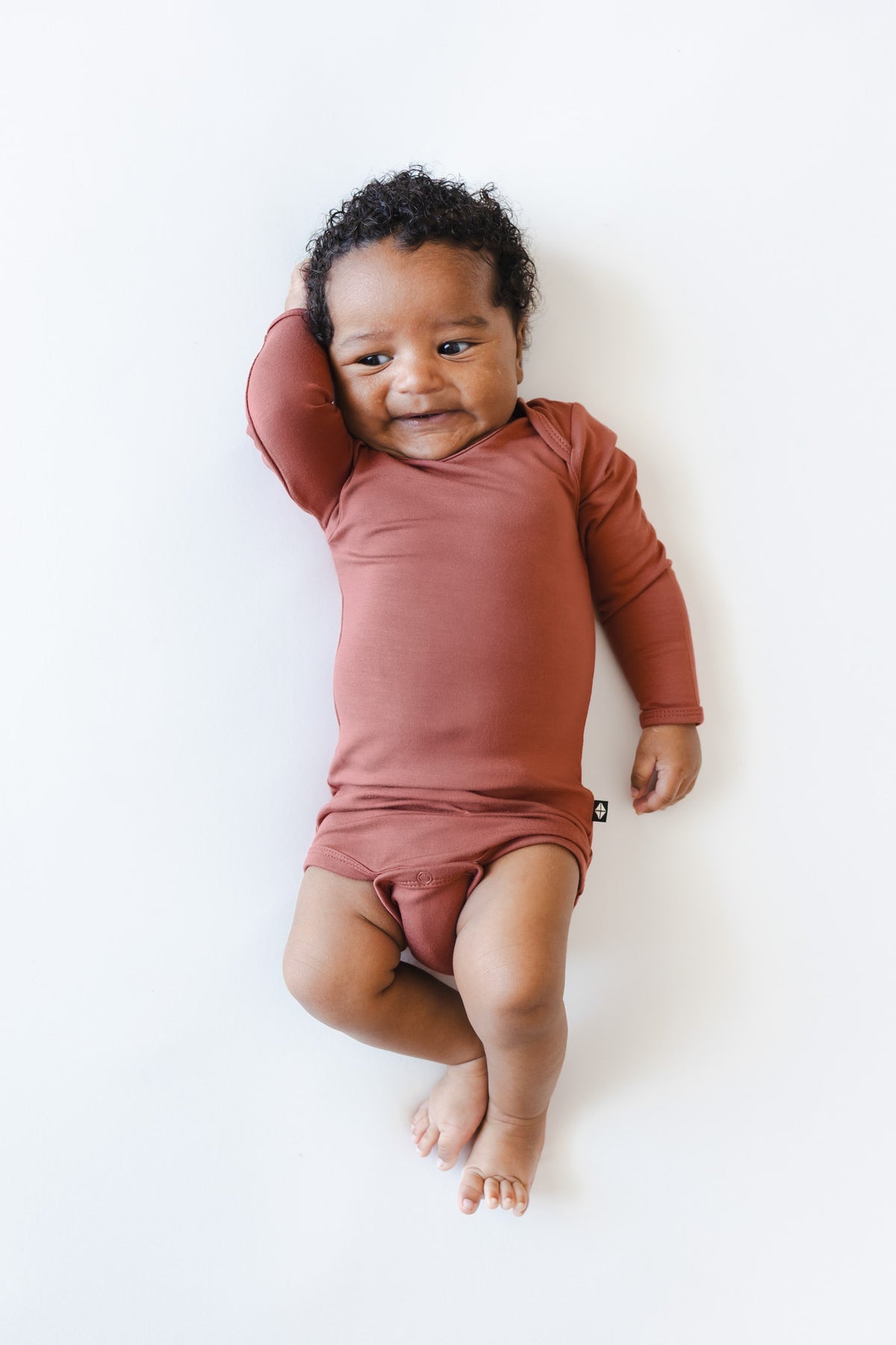 A baby lying on a white background wearing a dusty terracotta long-sleeve bodysuit.