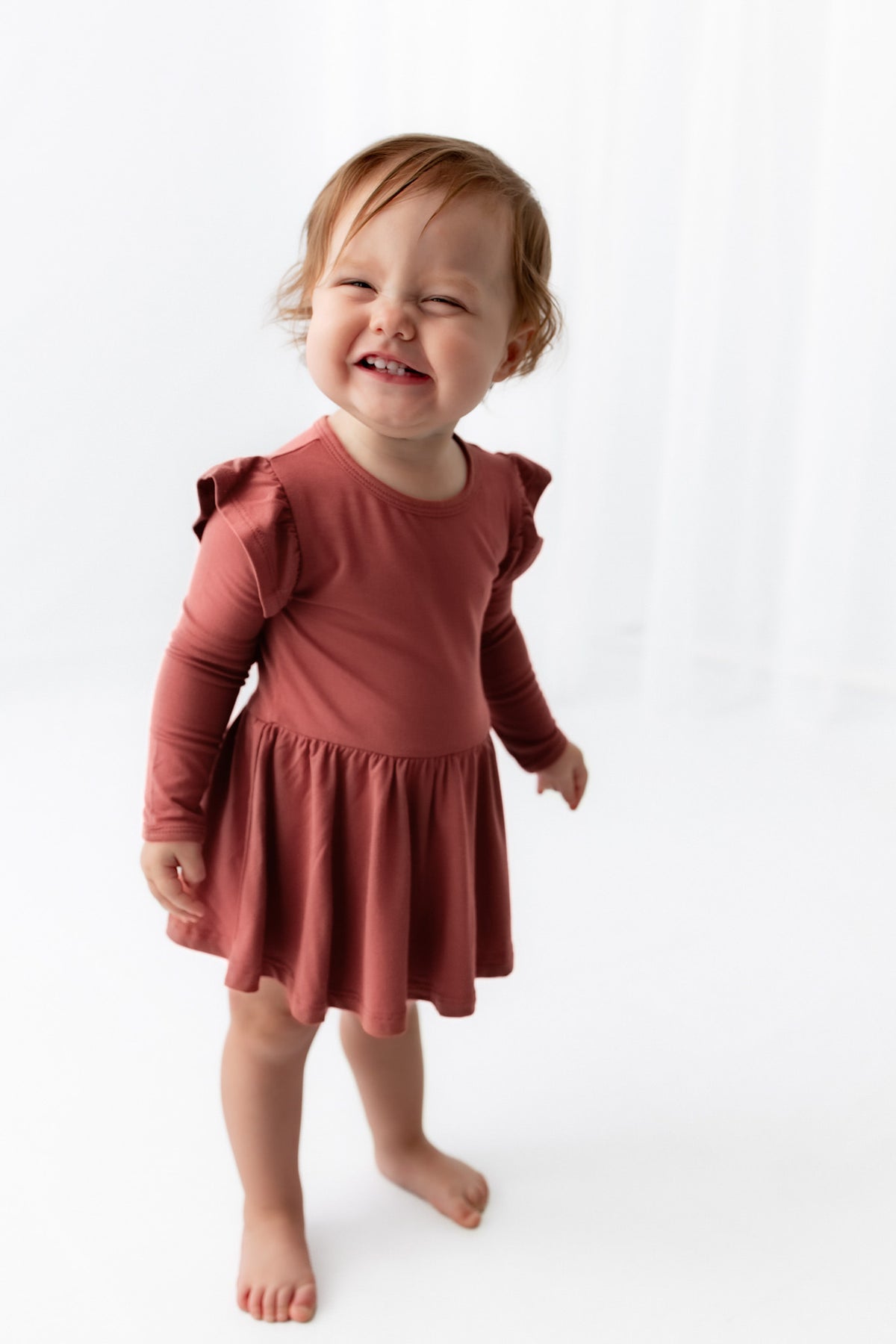 A toddler girl wearing a terracotta long-sleeve dress with ruffled shoulder details, standing barefoot against a clean white studio background.