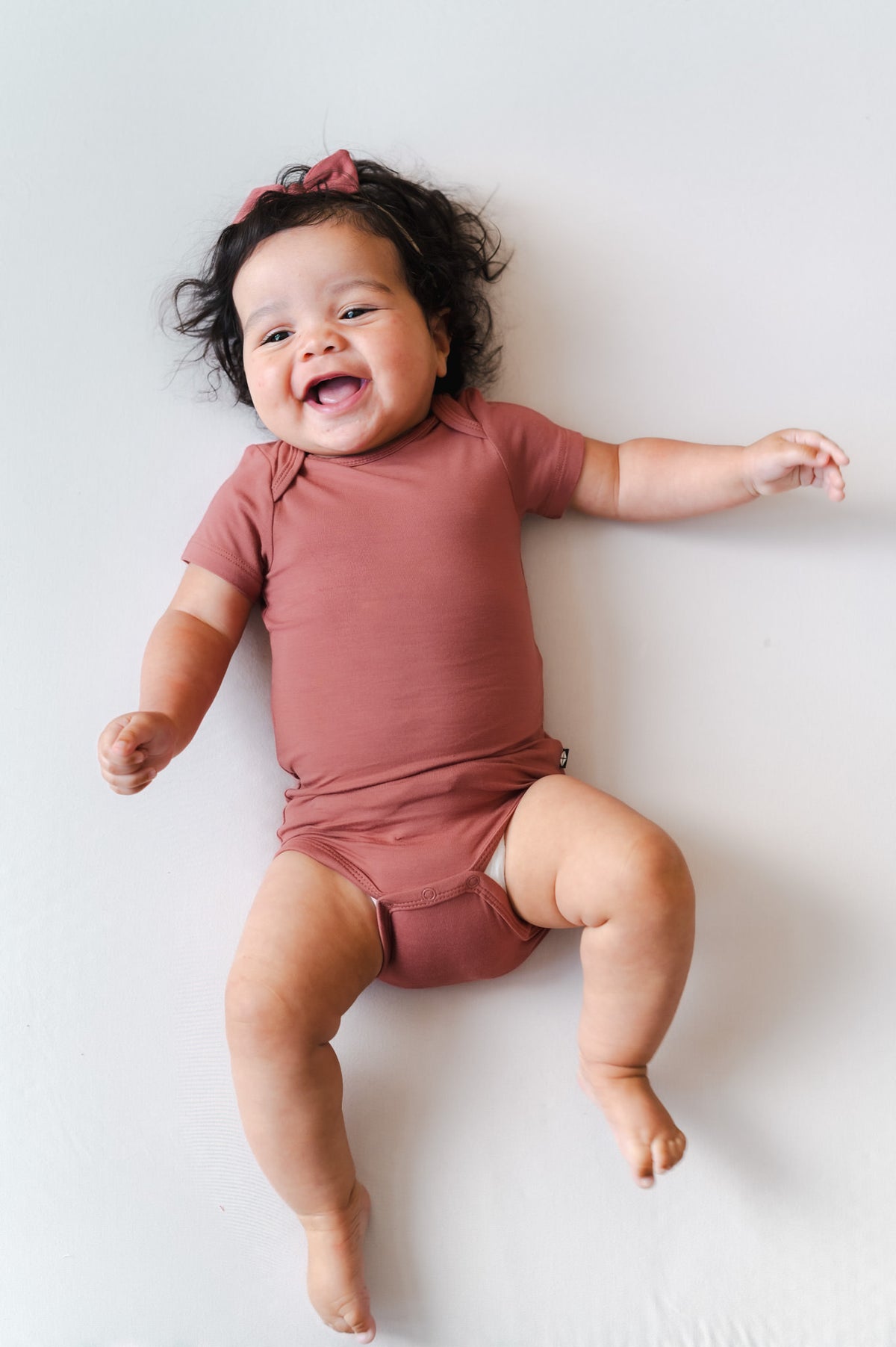 A smiling baby lying on their back against a plain white background, wearing a dusty terracotta short-sleeve bodysuit and a matching hair bow.