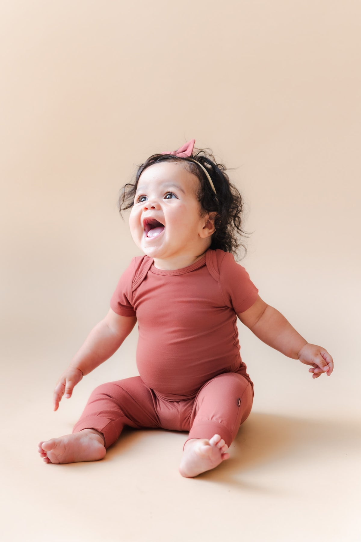 A smiling baby sitting on a warm beige studio background, wearing a matching dusty terracotta short-sleeve bodysuit and a small pink bow headband.