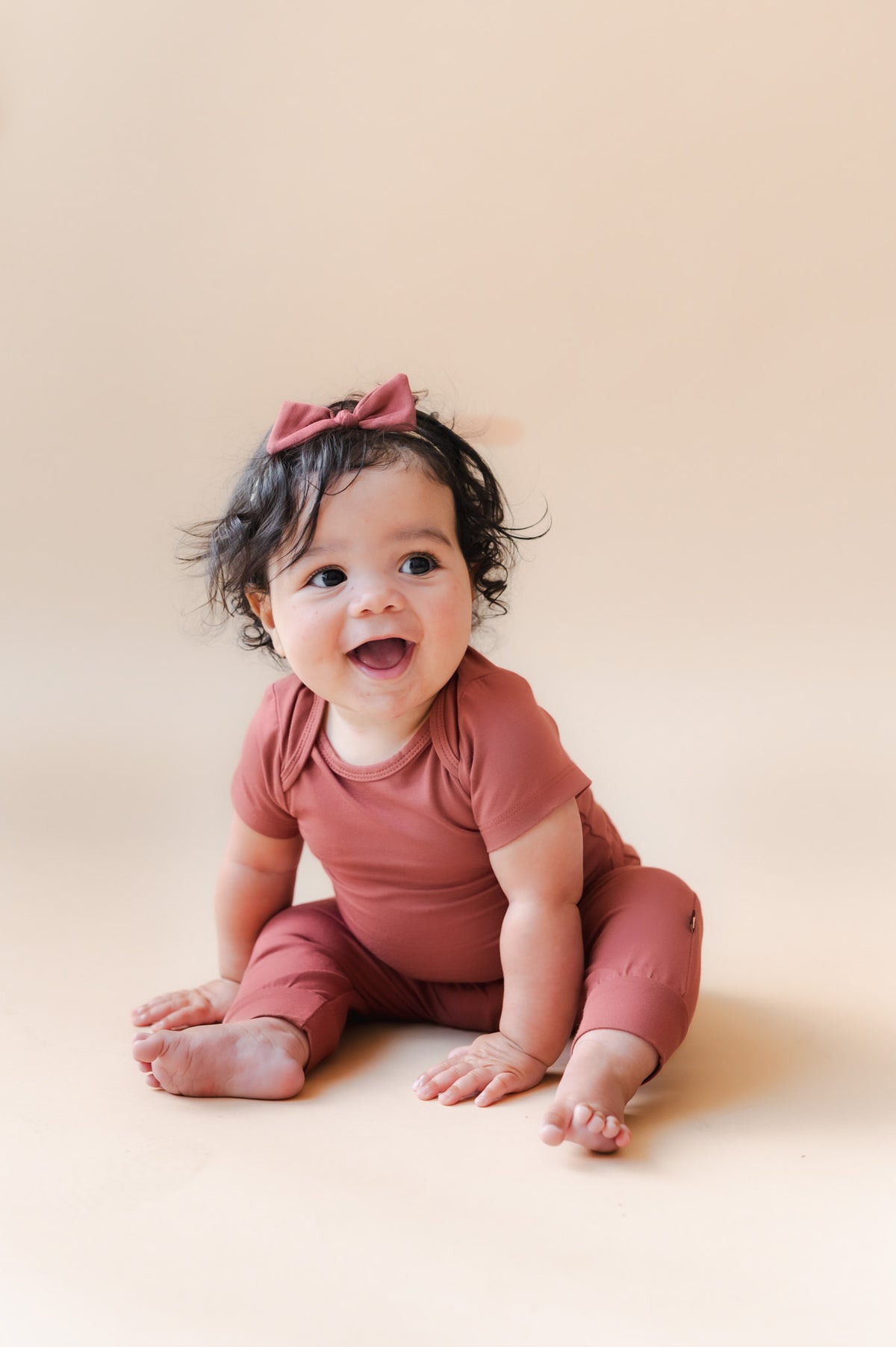 A smiling baby sitting on a warm beige studio background, wearing a matching dusty terracotta short-sleeve bodysuit and matching bow headband.