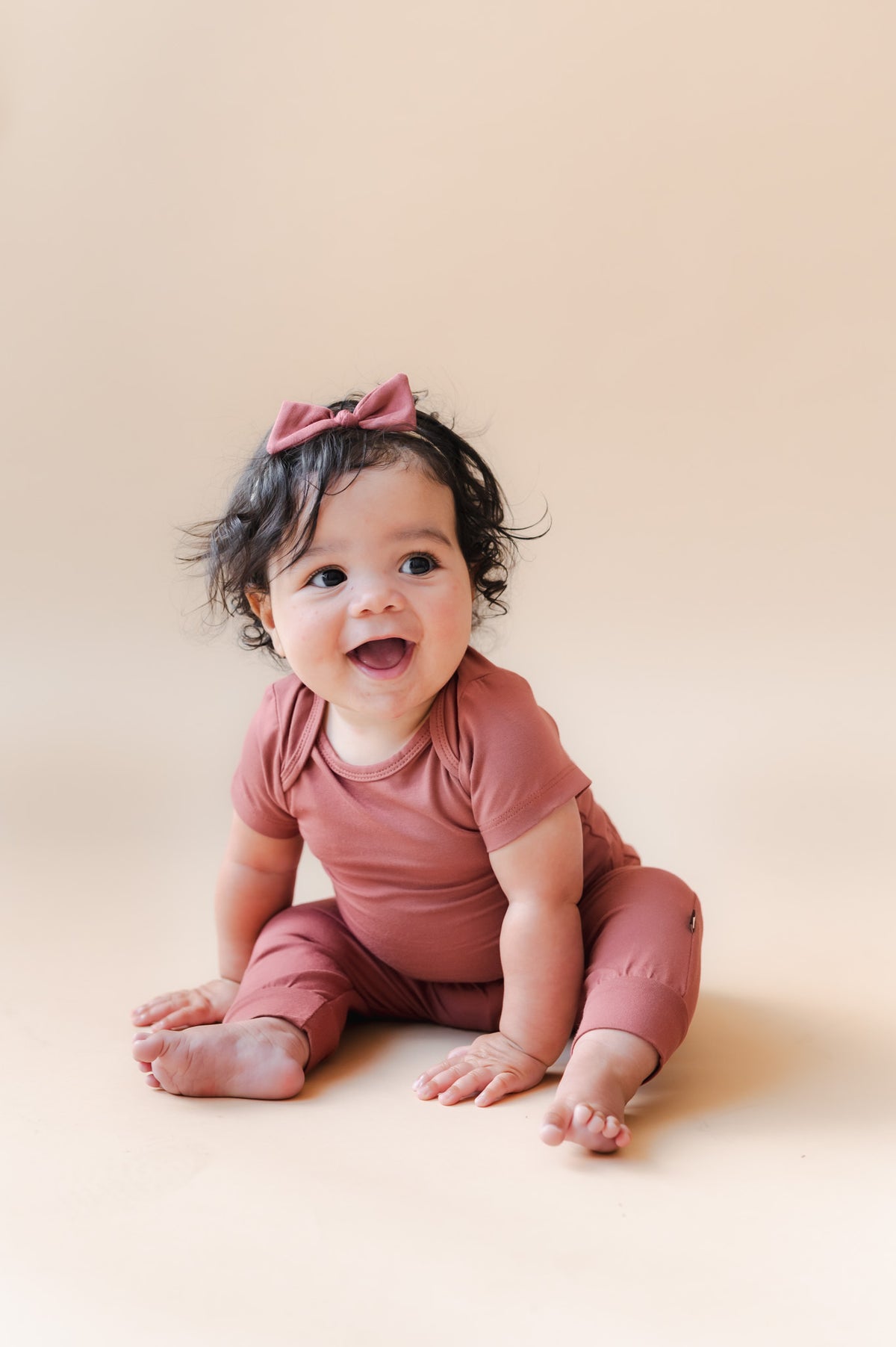 Baby sitting wearing Pant in Ember over similar color bodysuit and boy headband on a beige background