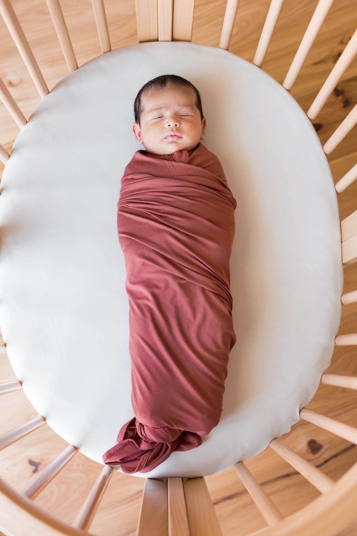 A sleeping newborn baby swaddled in a dusty terracotta blanket, lying on a white oval mattress inside a light wood slatted crib.