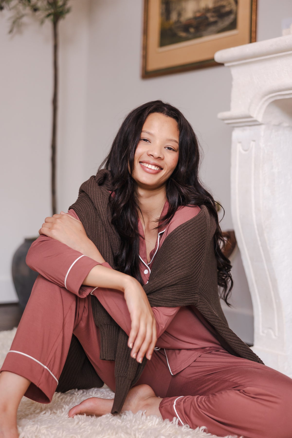 Modal smiling in Long-Sleeved Women's Pajama Set in Ember sitting on floor with a brown sweater over her shoulders