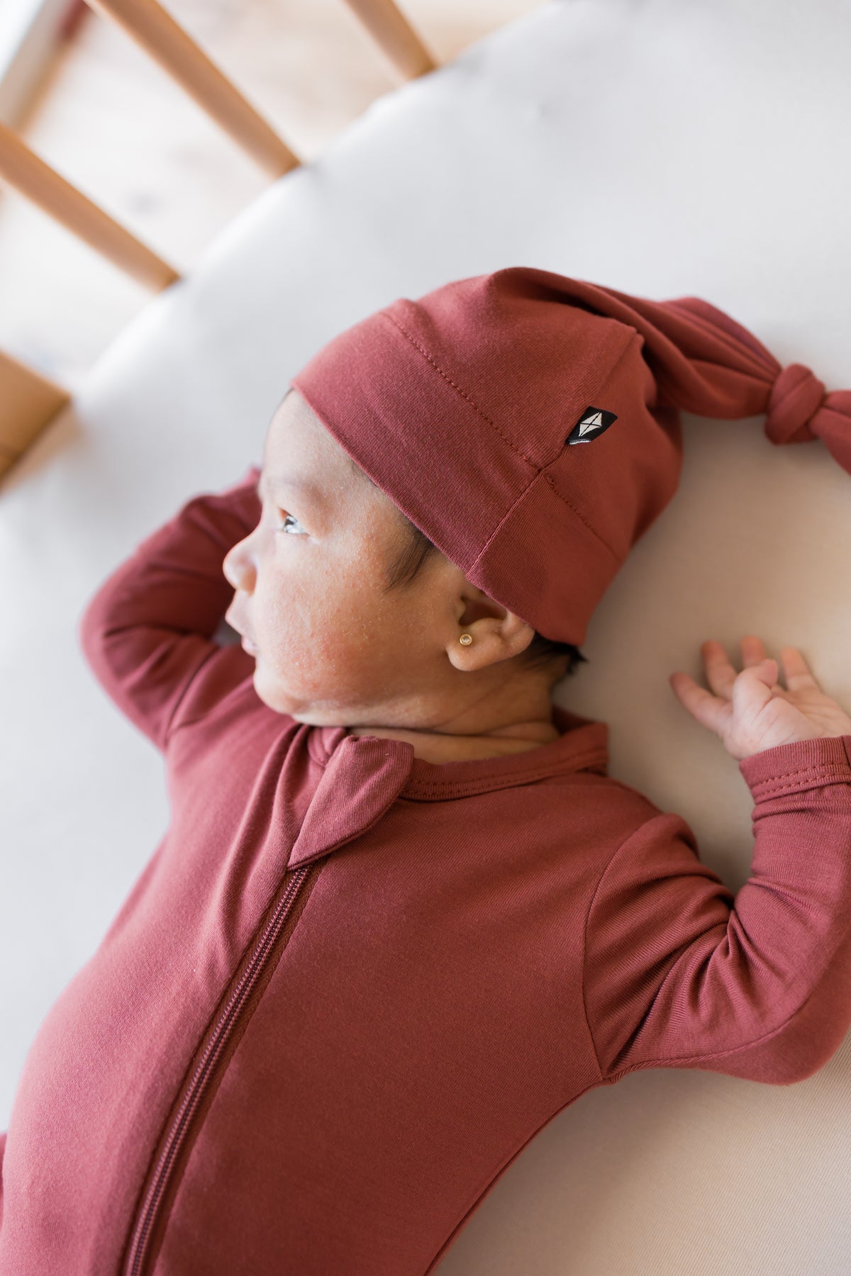 Baby wearing Knotted Cap in Ember with zippered footie in ember lying in a crib