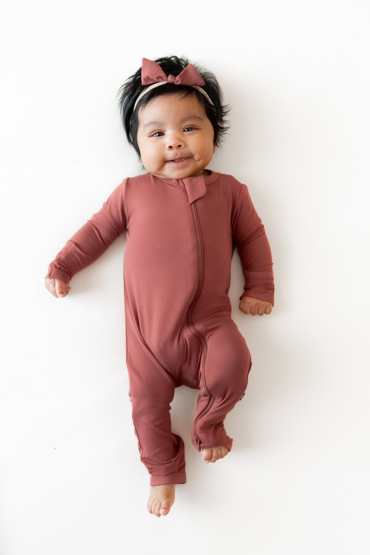 A baby lying on a white background wearing a dusty terracotta long-sleeve footless one-piece romper and a matching bow headband.