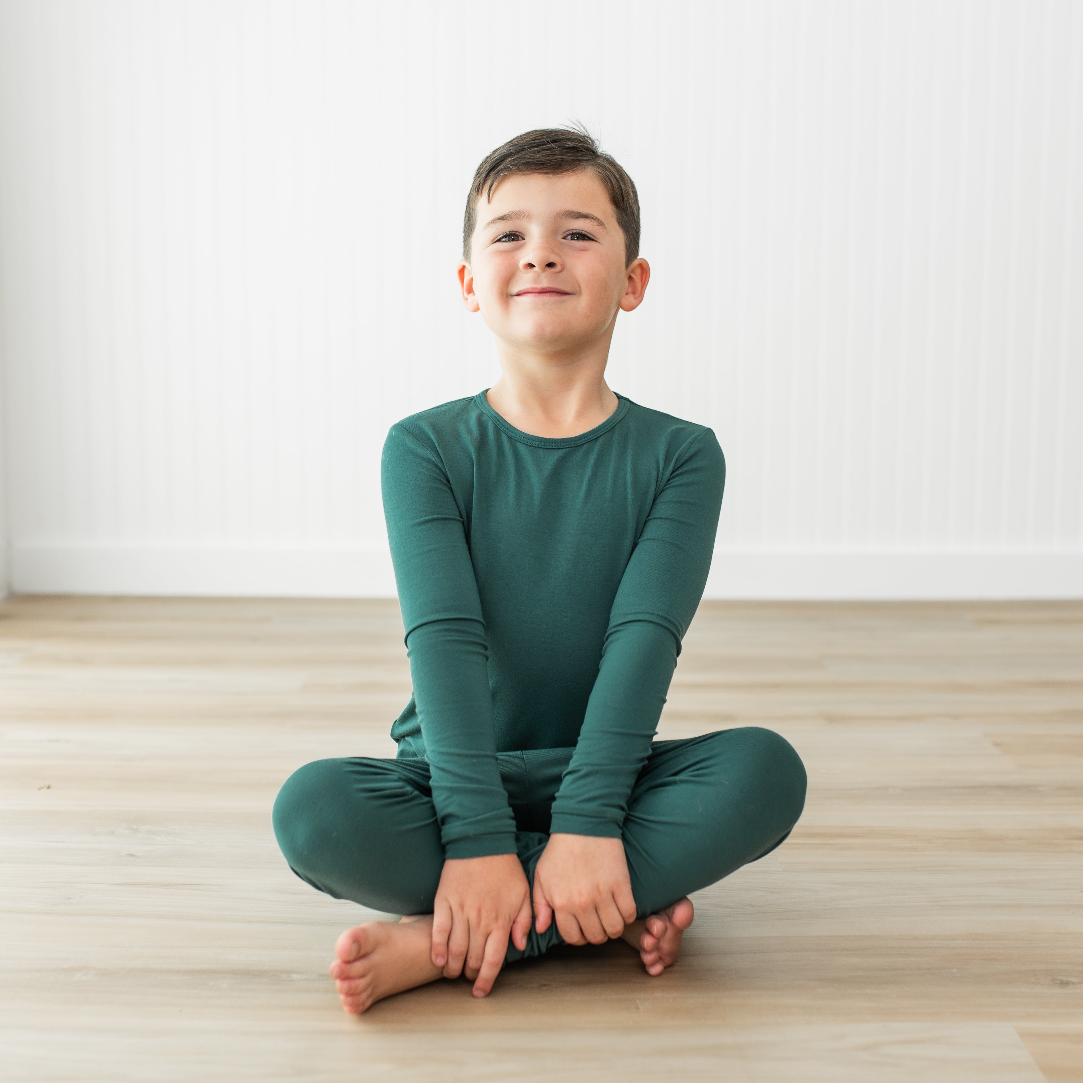 Young boy sitting cross-legged on a wooden floor wearing a green outfit.