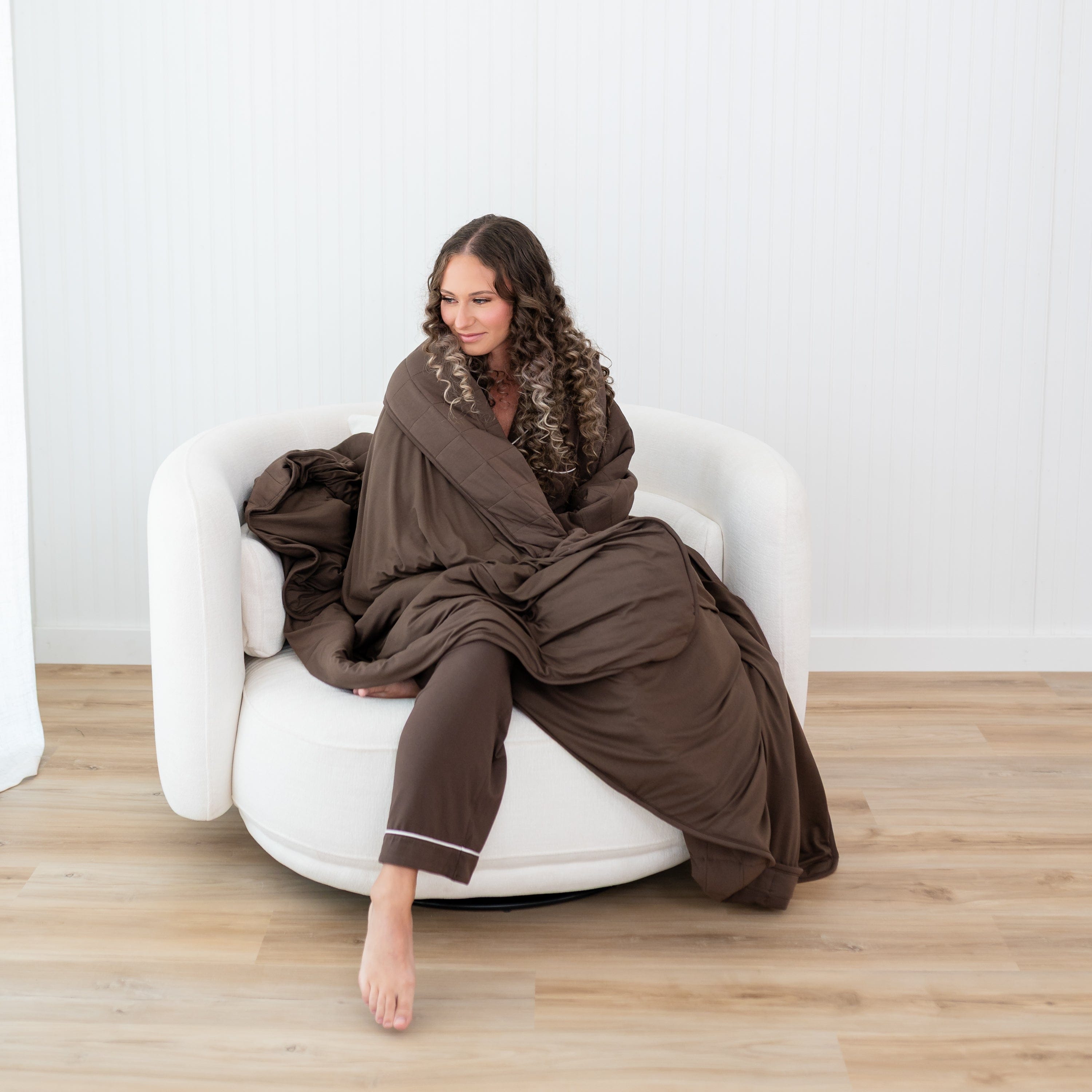 Female model sitting on a couch with the Adult Quilted Blanket in Espresso 2.5 wrapped around her