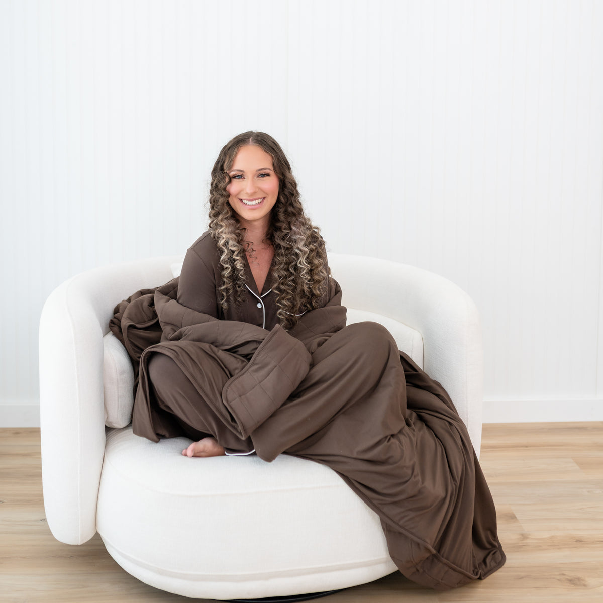 Smiling female model sitting on a couch with the Adult Quilted Blanket in Espresso 2.5 wrapped around her