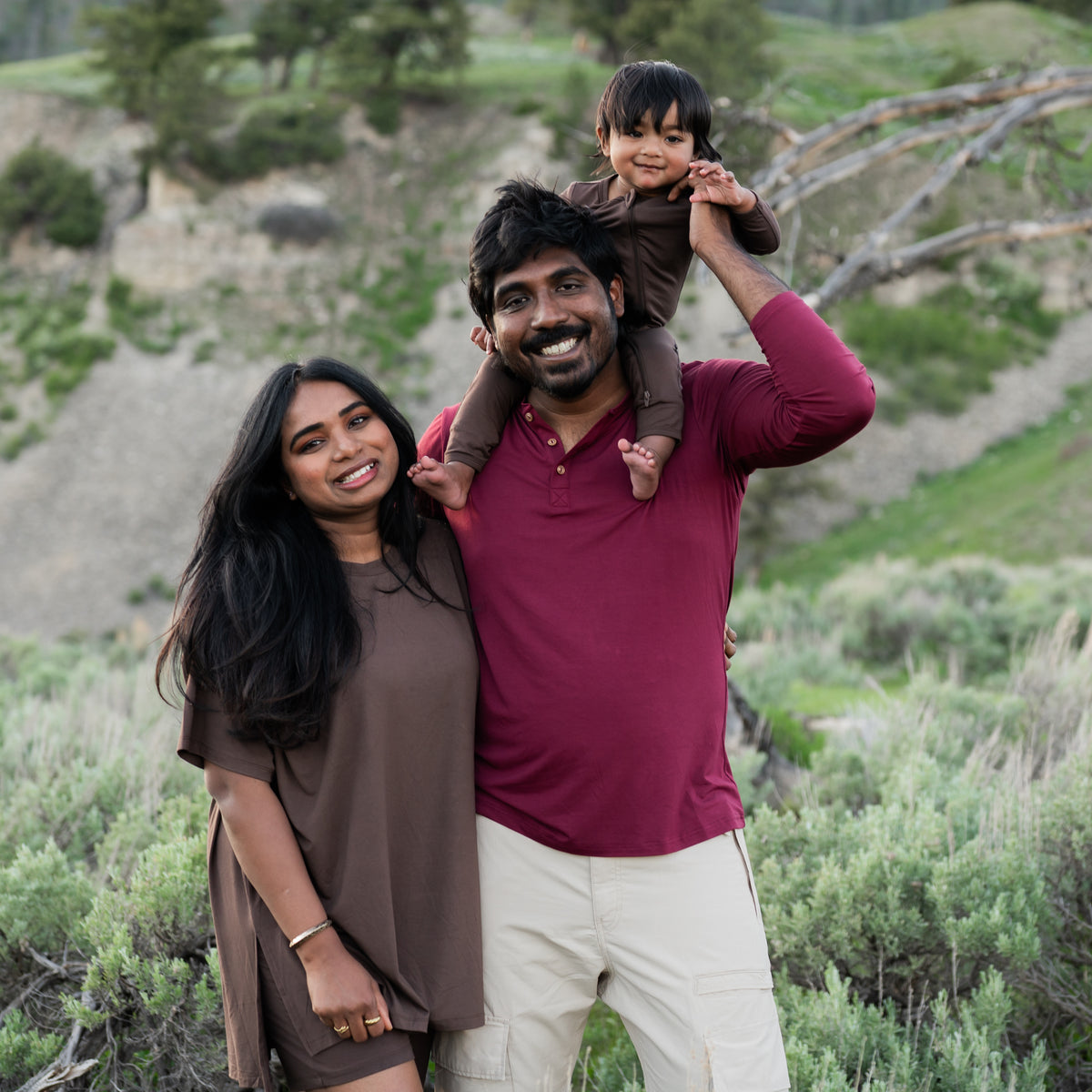 Family standing together outside in a field. Toddler is waring the Espresso zippered romper sitting on the dads shoulder. Dad is wearing the Men's Long Sleeve Henley Top in Burgundy while Mom is wearing the Espresso Women's Biker short set