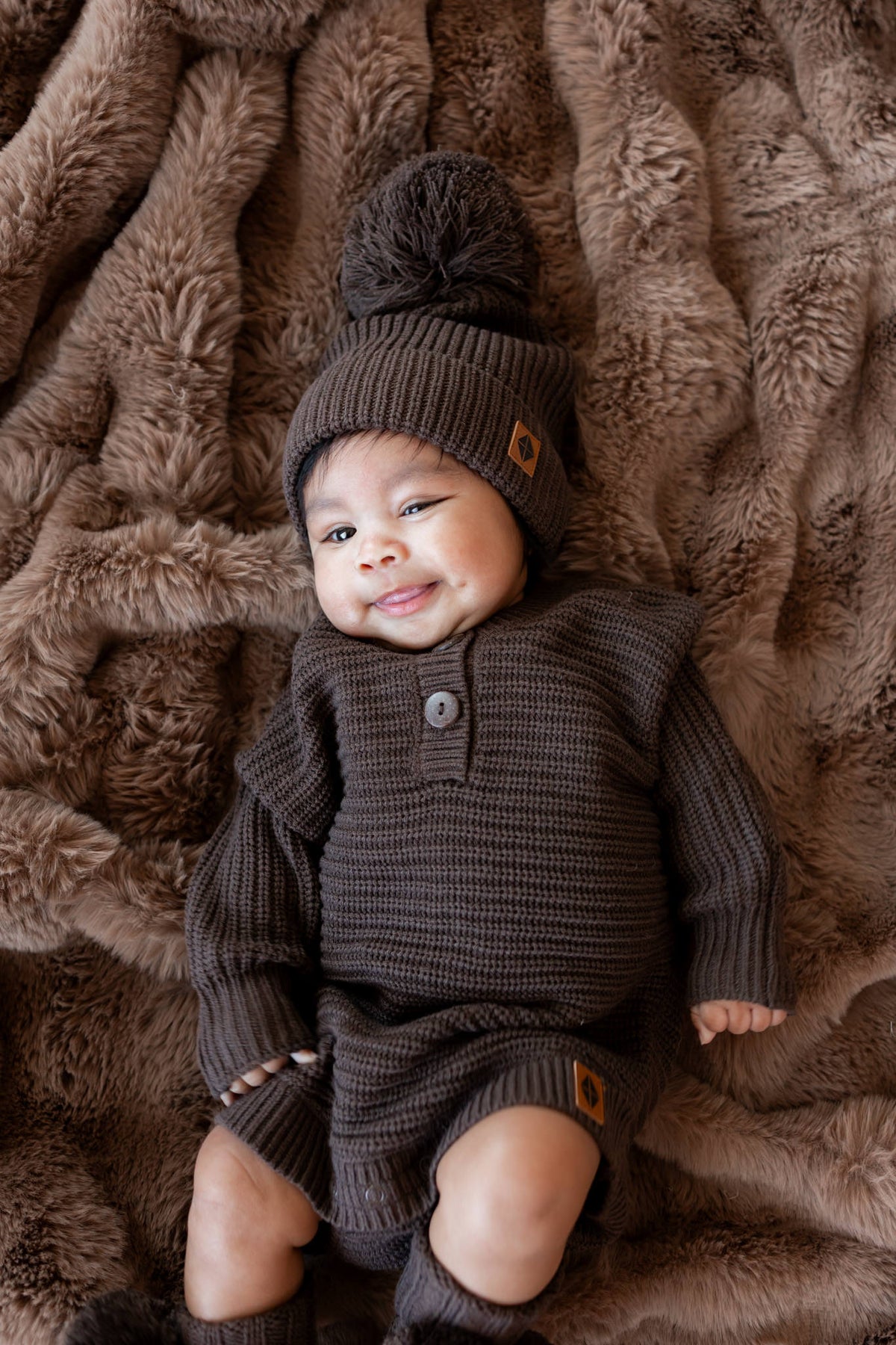 Baby wearing wearing Chunky knit romper, Chunky knit pom beanie and knit booties in Espresso on a brown blanket