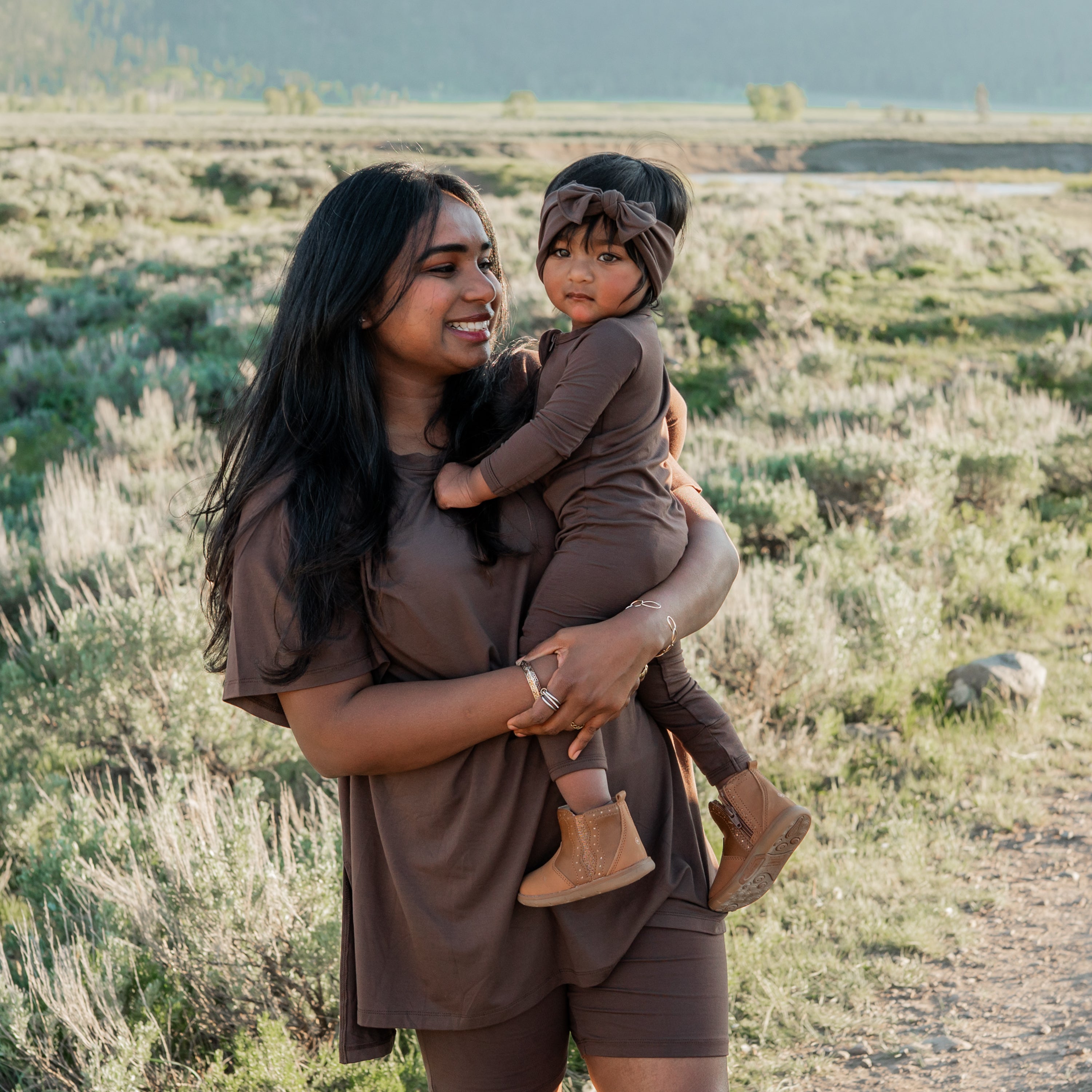 Mother and daughter matching in Espresso. Mother wearing the Women’s Biker Short Set in Espresso holding her daughter wearing the Zippered romper and knotted bow headband