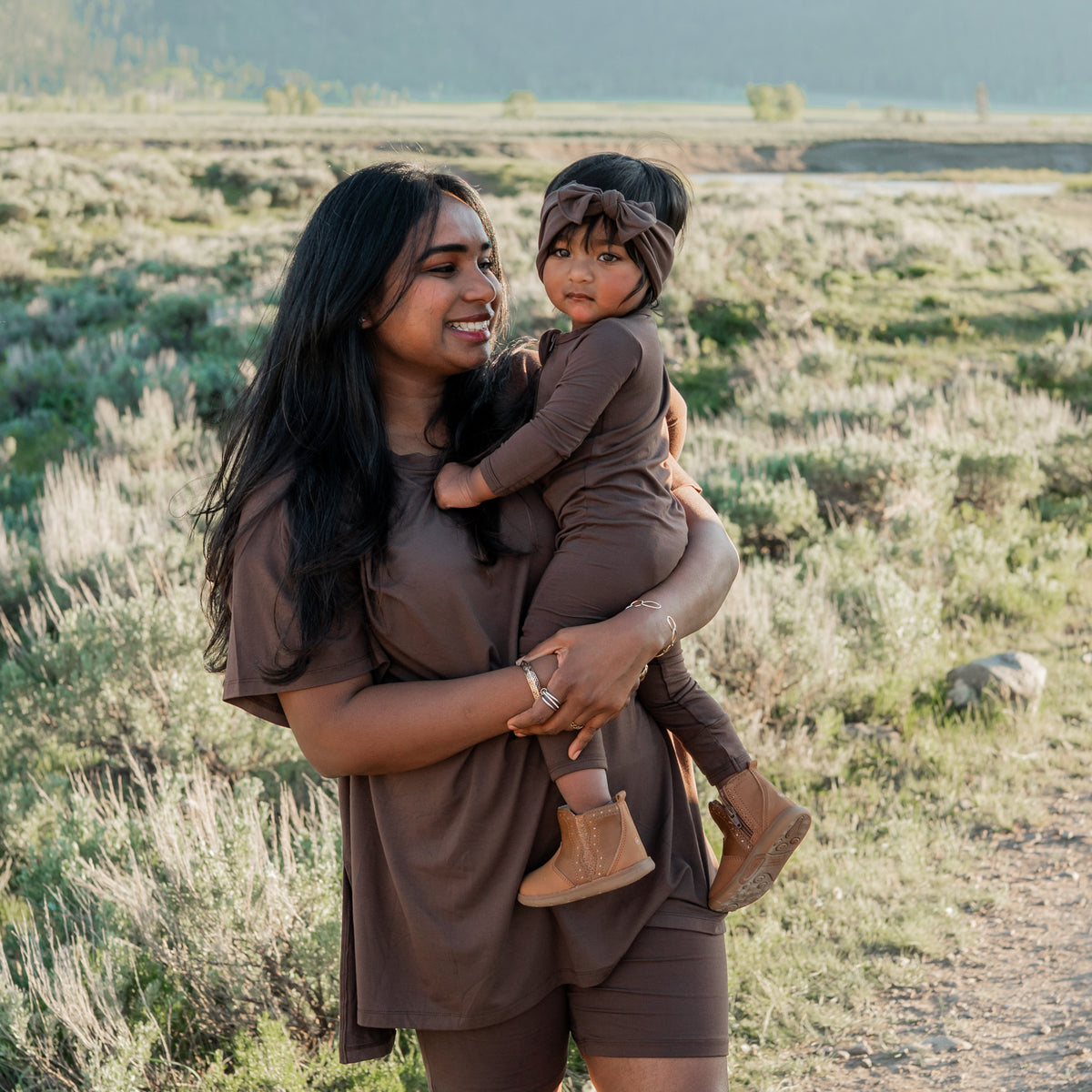 Mother and daughter matching in Espresso. Mother wearing the Women’s Biker Short Set in Espresso holding her daughter wearing the Zippered romper and knotted bow headband