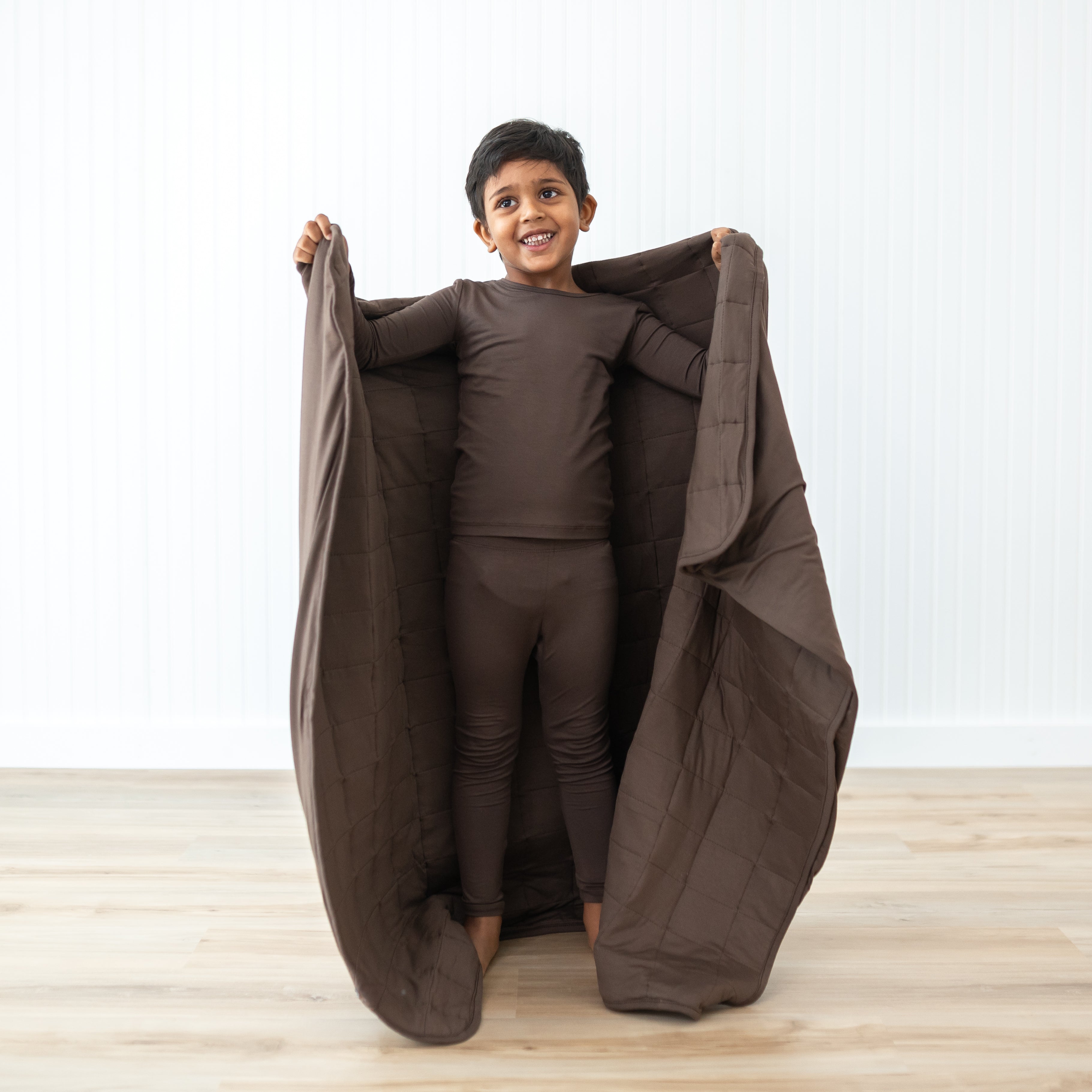 Smiling boy model standing holding the Toddler Blanket in Espresso 2.5 around him wearing matching toddler pajamas