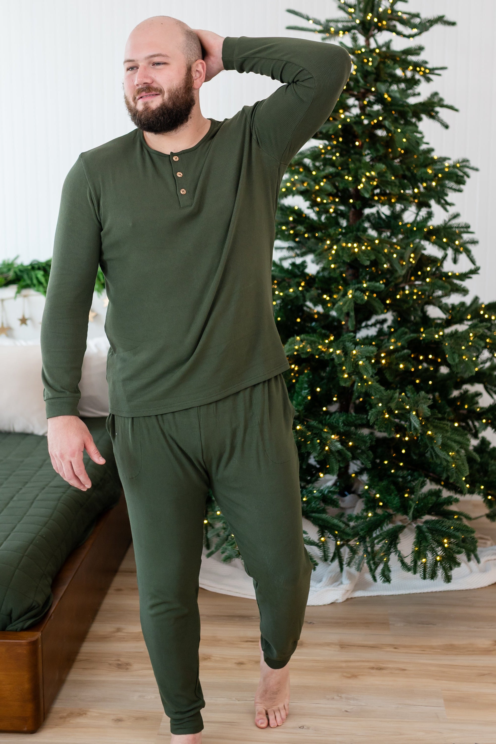 Male mode walking with one hand up behind his head wearing the Men's Ribbed Henley Set in Fir beside a bed and in front of a lit Christmas tree
