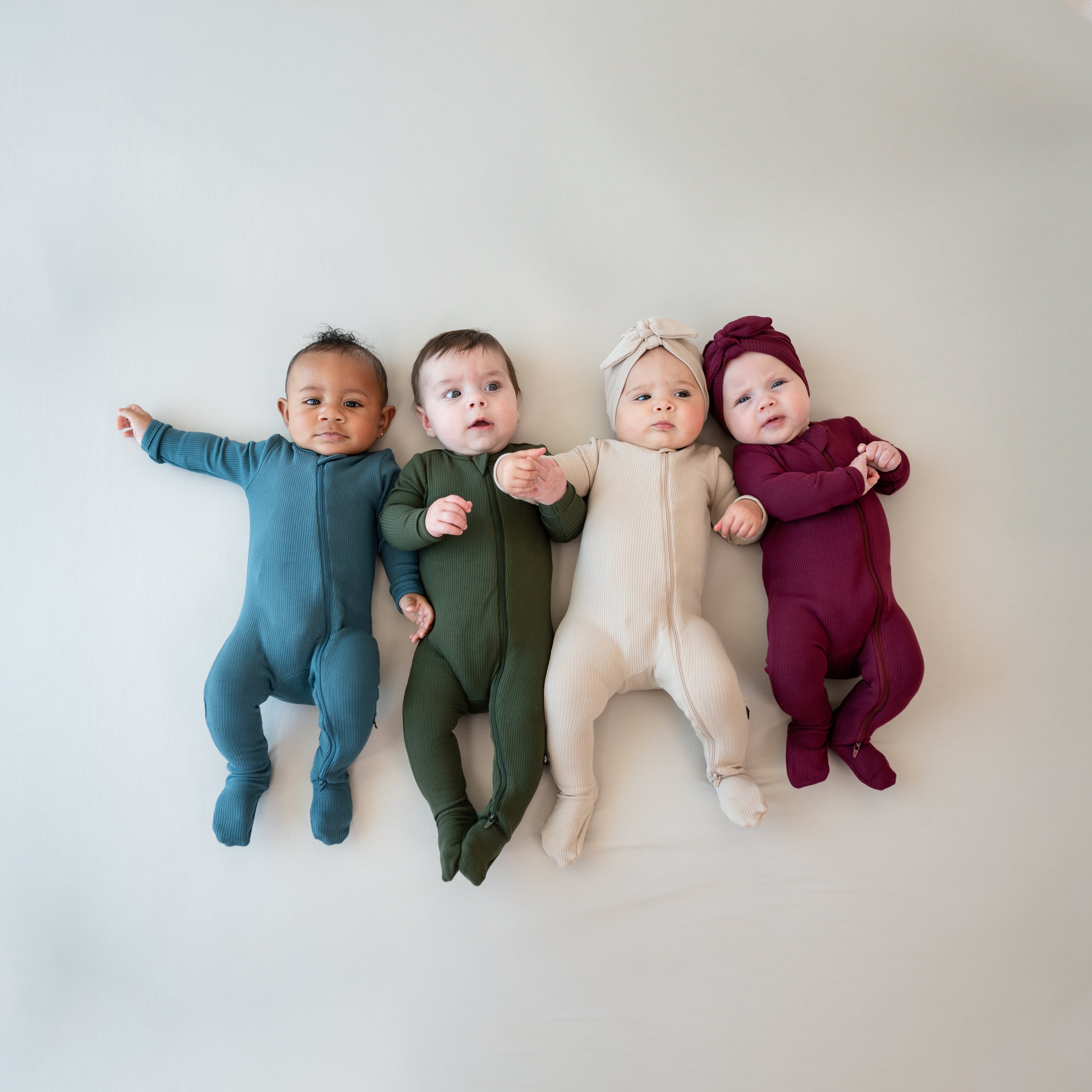 Four infants laying side by side wearing the Ribbed Zipper Footies in Bisque, Burgundy, Fir and Atlantic