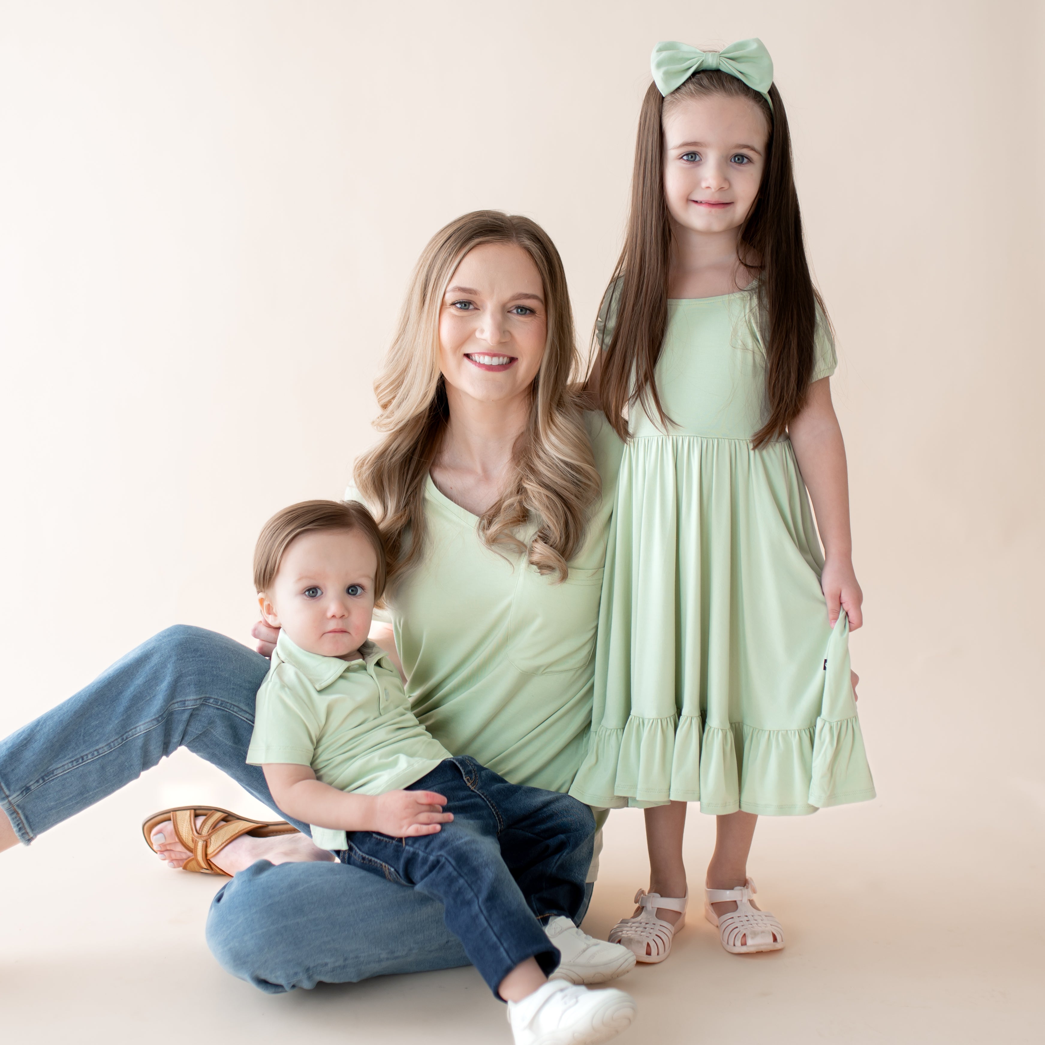 Woman with two children, one baby and one toddler, all wearing light green outfits on a plain background.