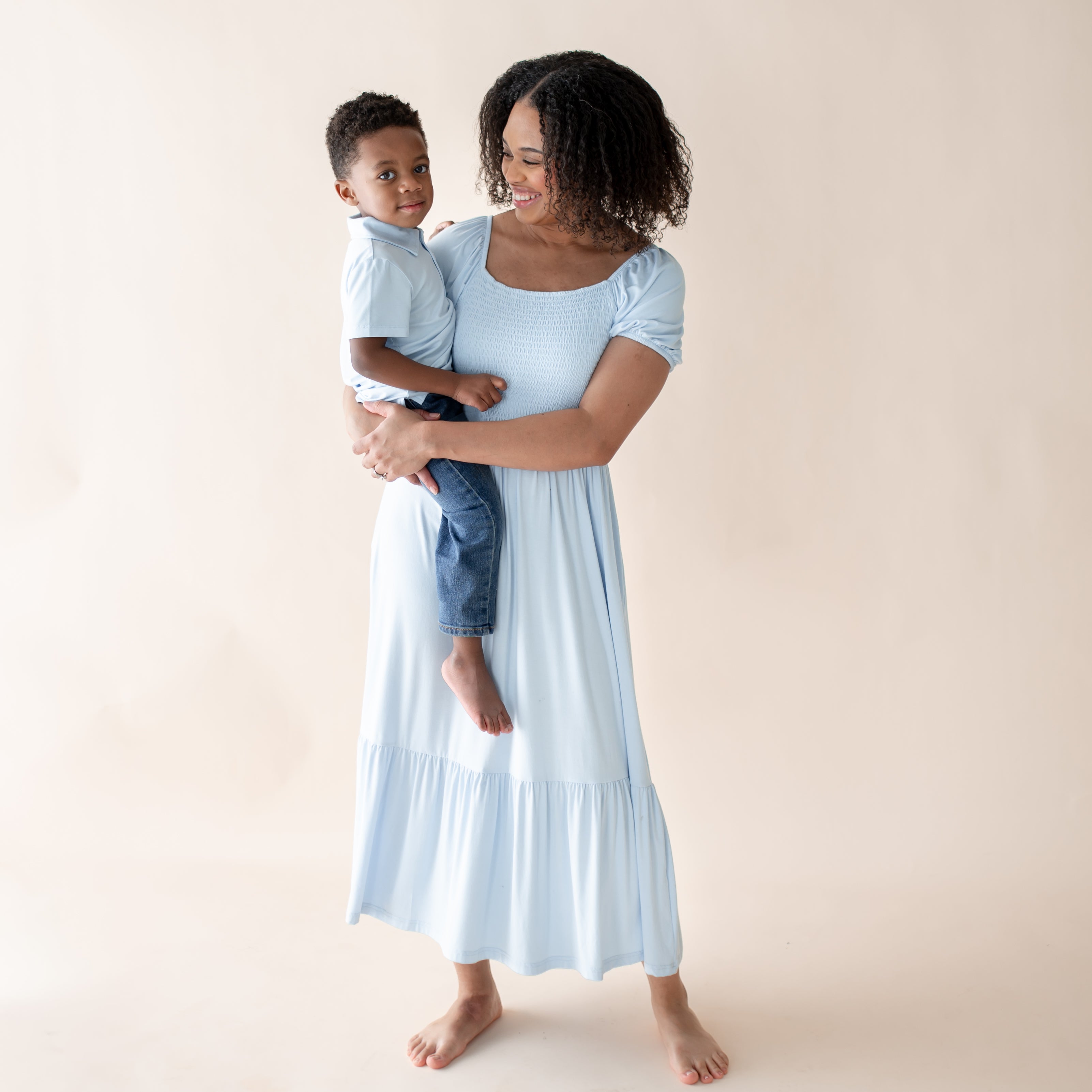Woman in a light blue dress holding a child in a matching blue polo against a beige background